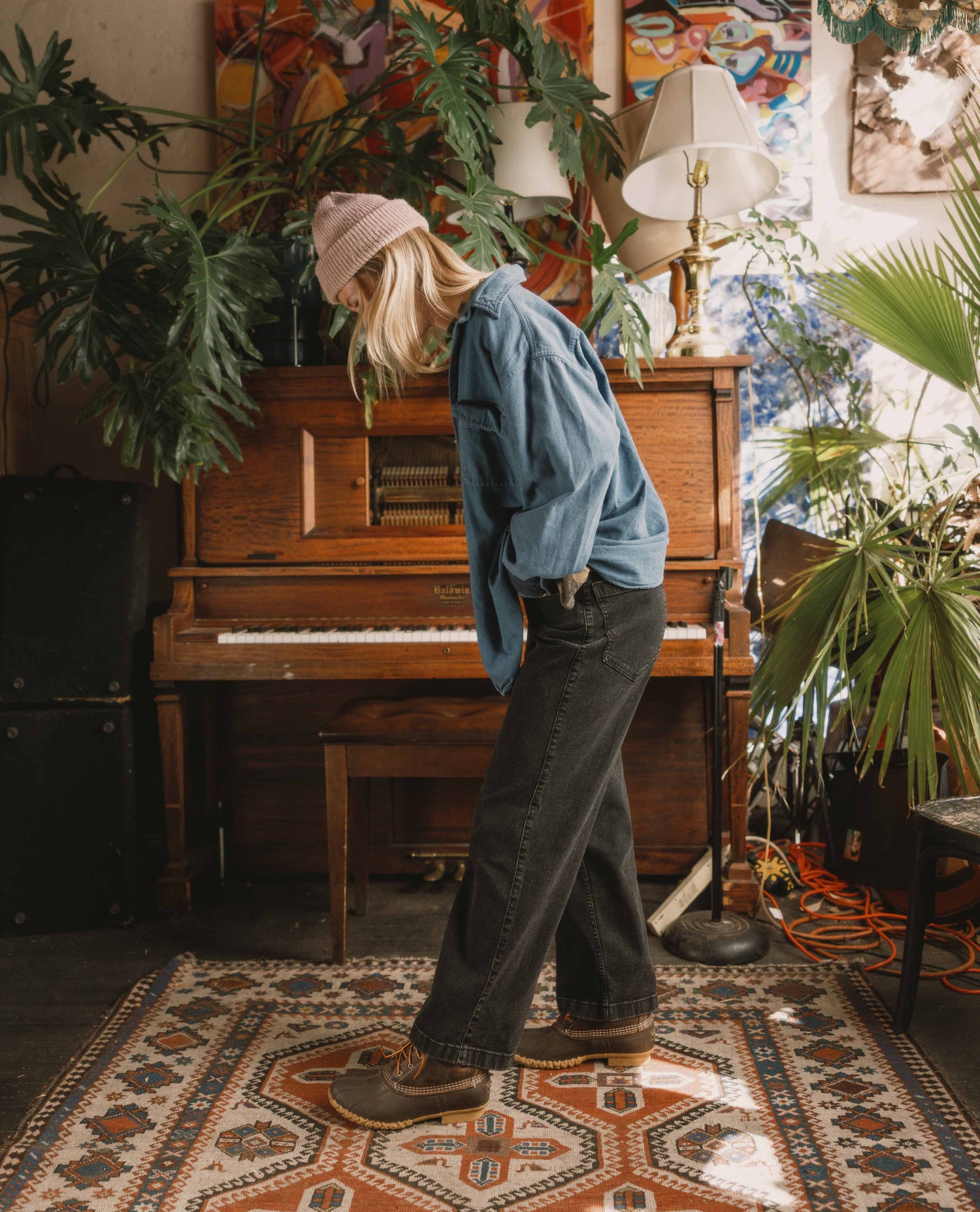 Person in a room with a piano and plants, wearing a blue shirt and brown shoes.
