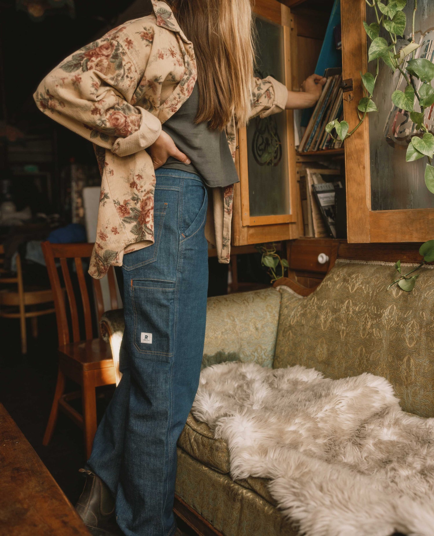 Person wearing a floral jacket and blue jeans in a cozy room with a couch and bookshelf.