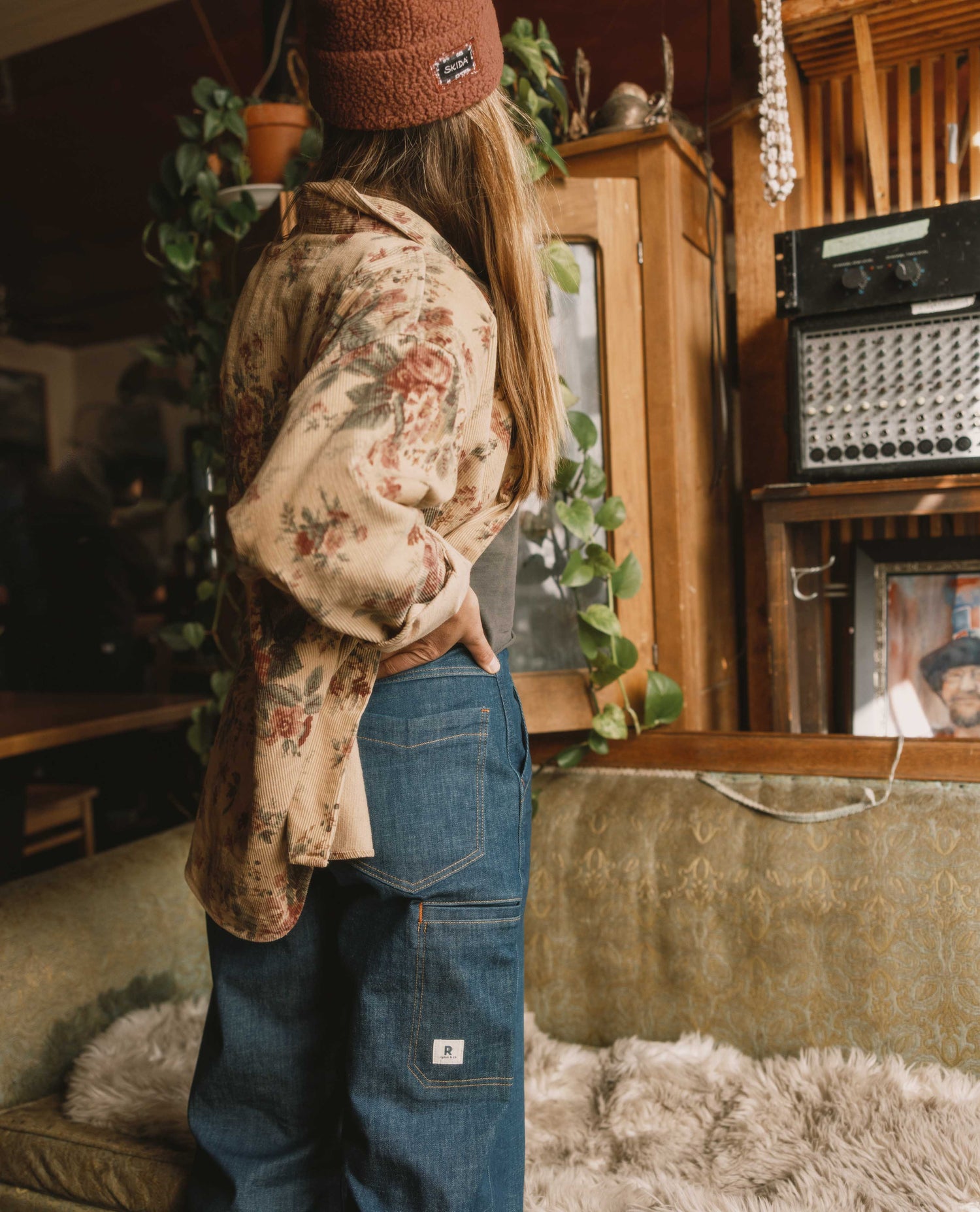 Person wearing a floral shirt and blue jeans in a cozy room with plants and vintage furniture.