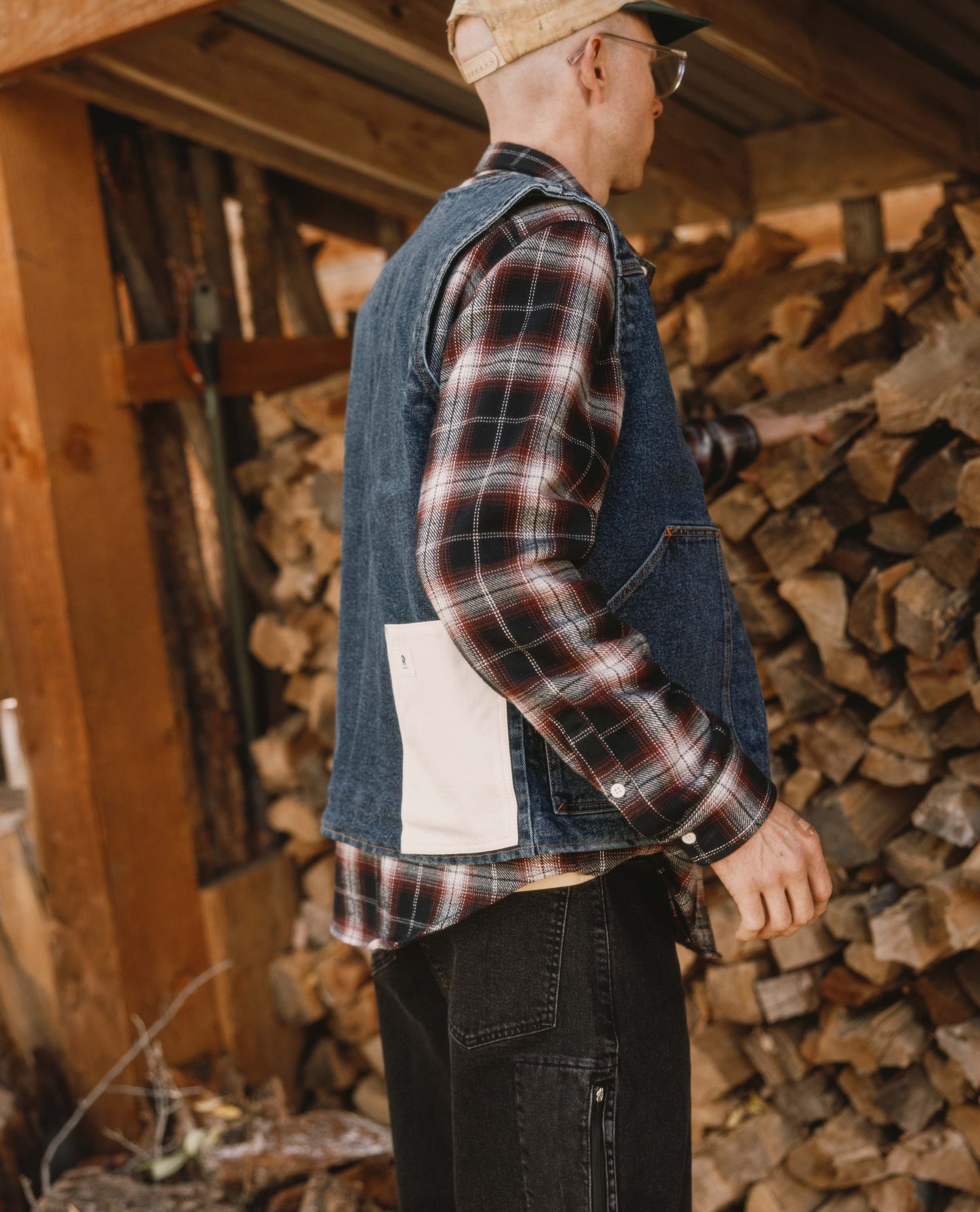 Man wearing a plaid shirt and denim vest standing next to stacked firewood.