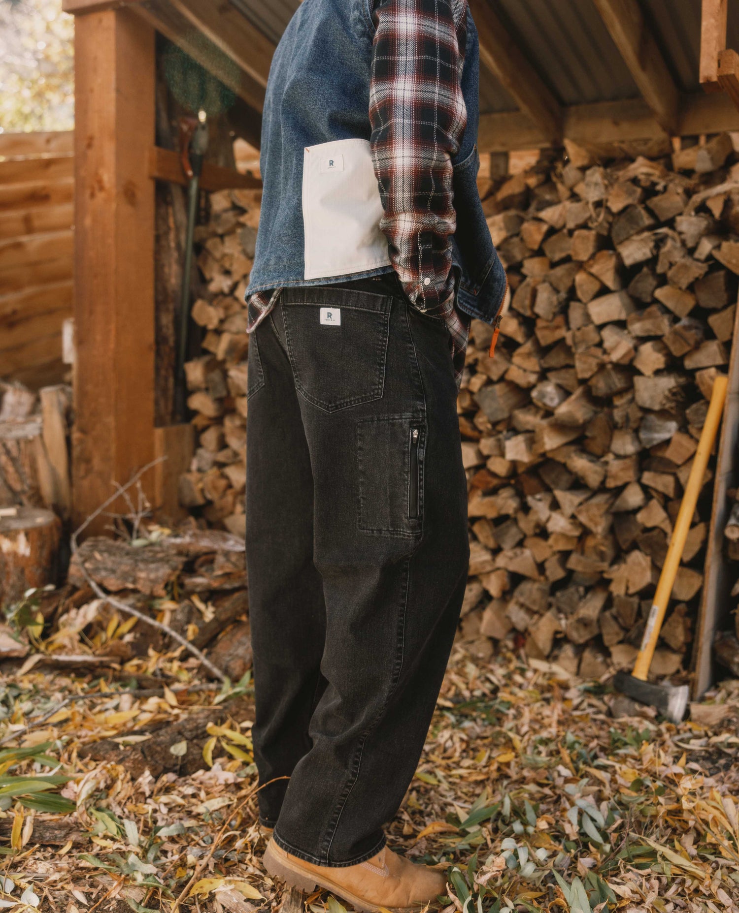 Person wearing a plaid shirt and dark jeans standing in front of stacked firewood.