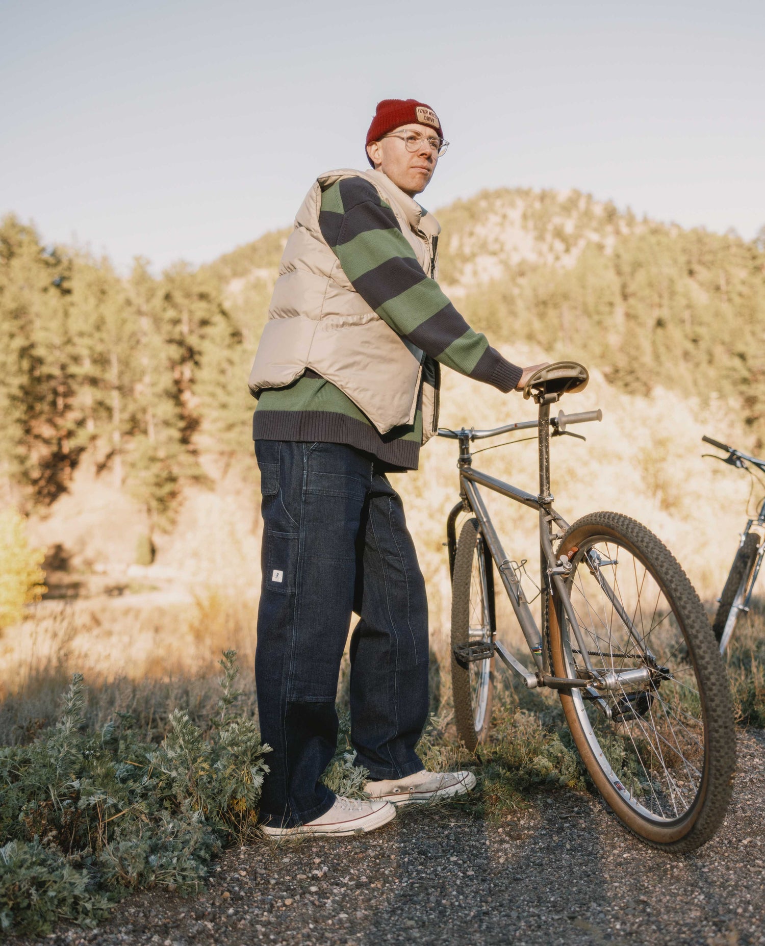 Person standing next to a bicycle in a natural setting with trees and open space.