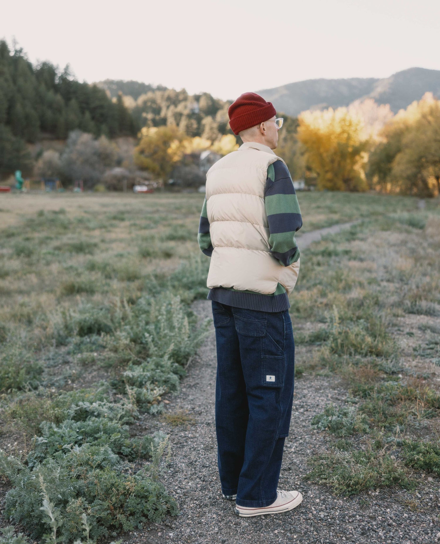 Person wearing a white puffer vest, red beanie, and navy pants standing on a path in a natural setting with trees and mountains.