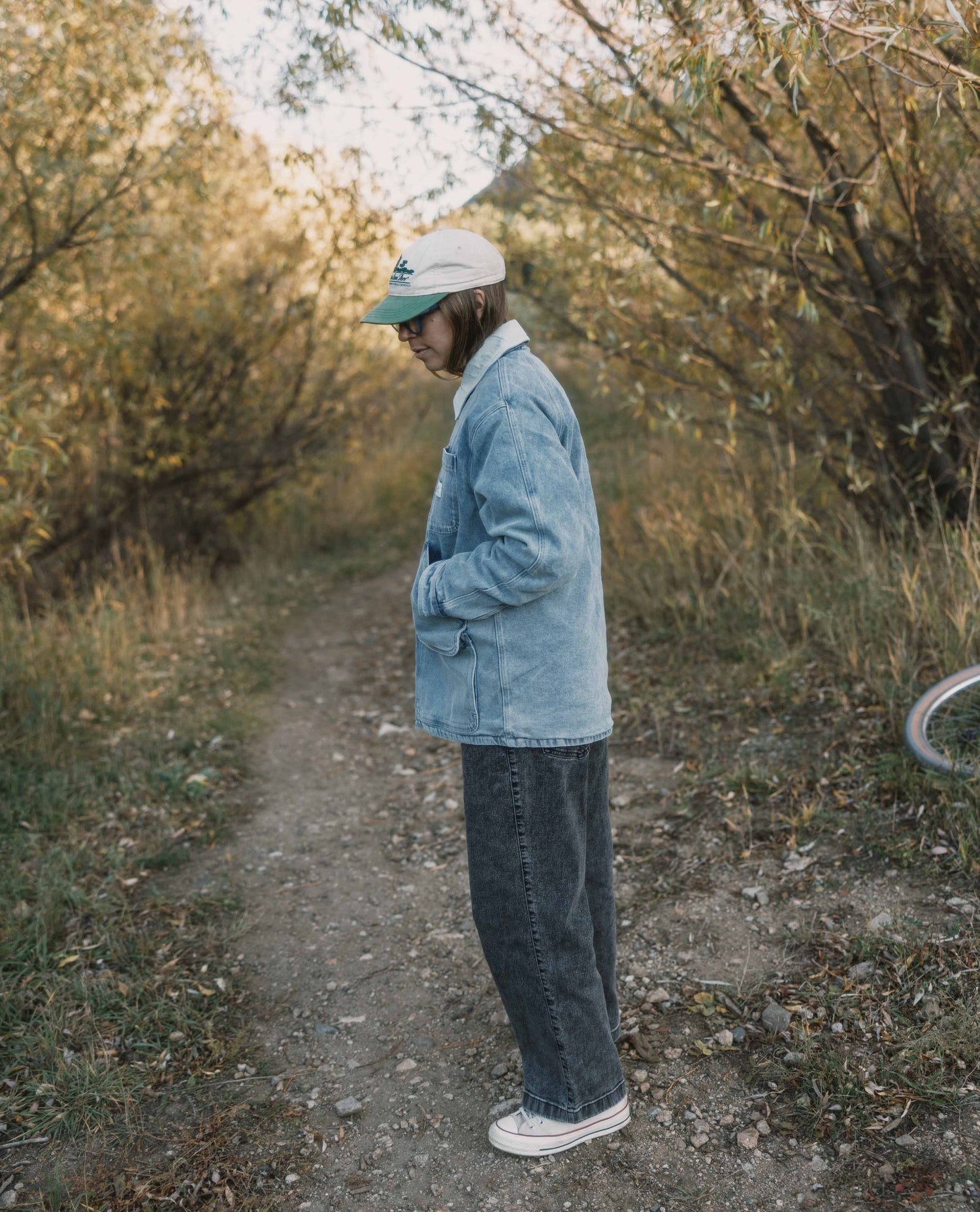 Person standing on a dirt path in a natural setting wearing a denim jacket and cap.