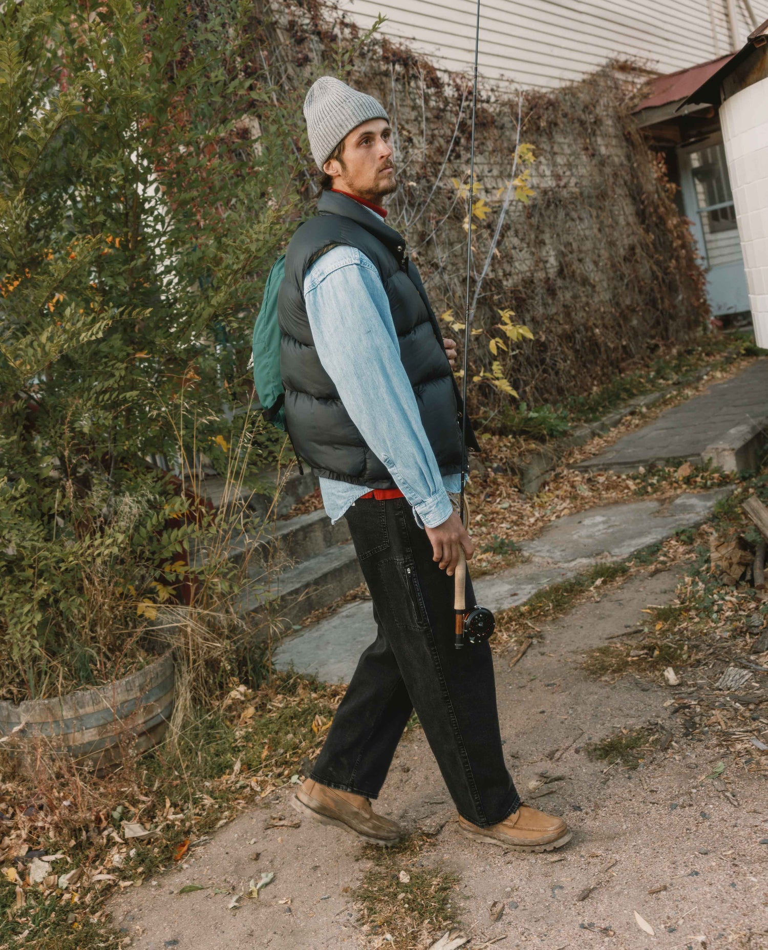 Man walking outdoors holding a rifle, wearing a vest and hat.