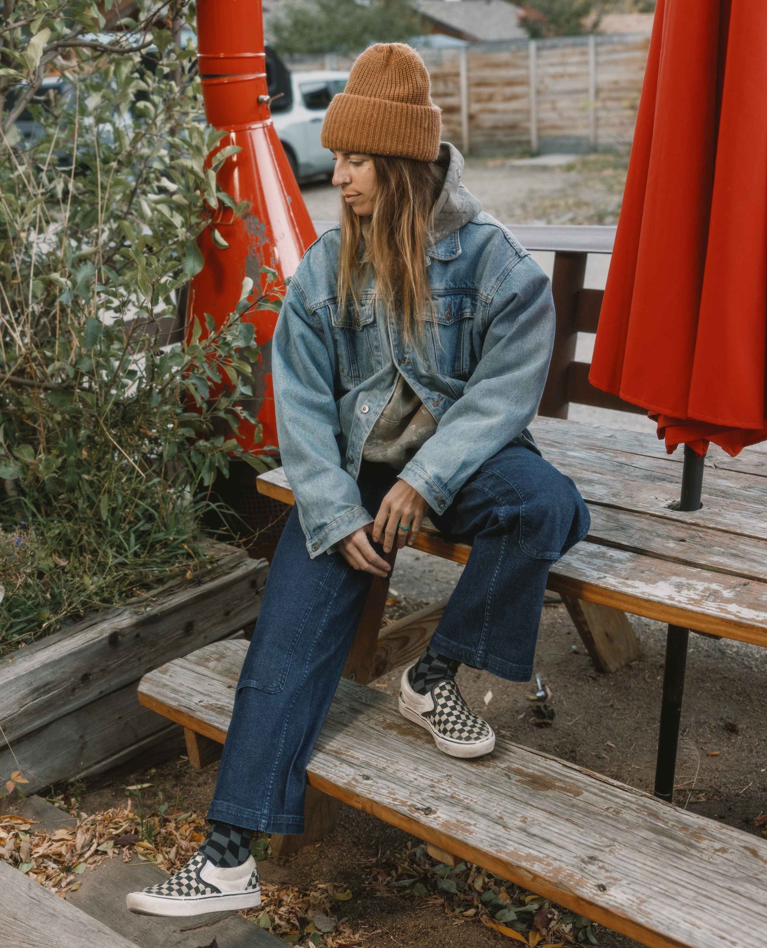 Person wearing a denim jacket and checkered shoes sitting on a wooden bench outdoors.