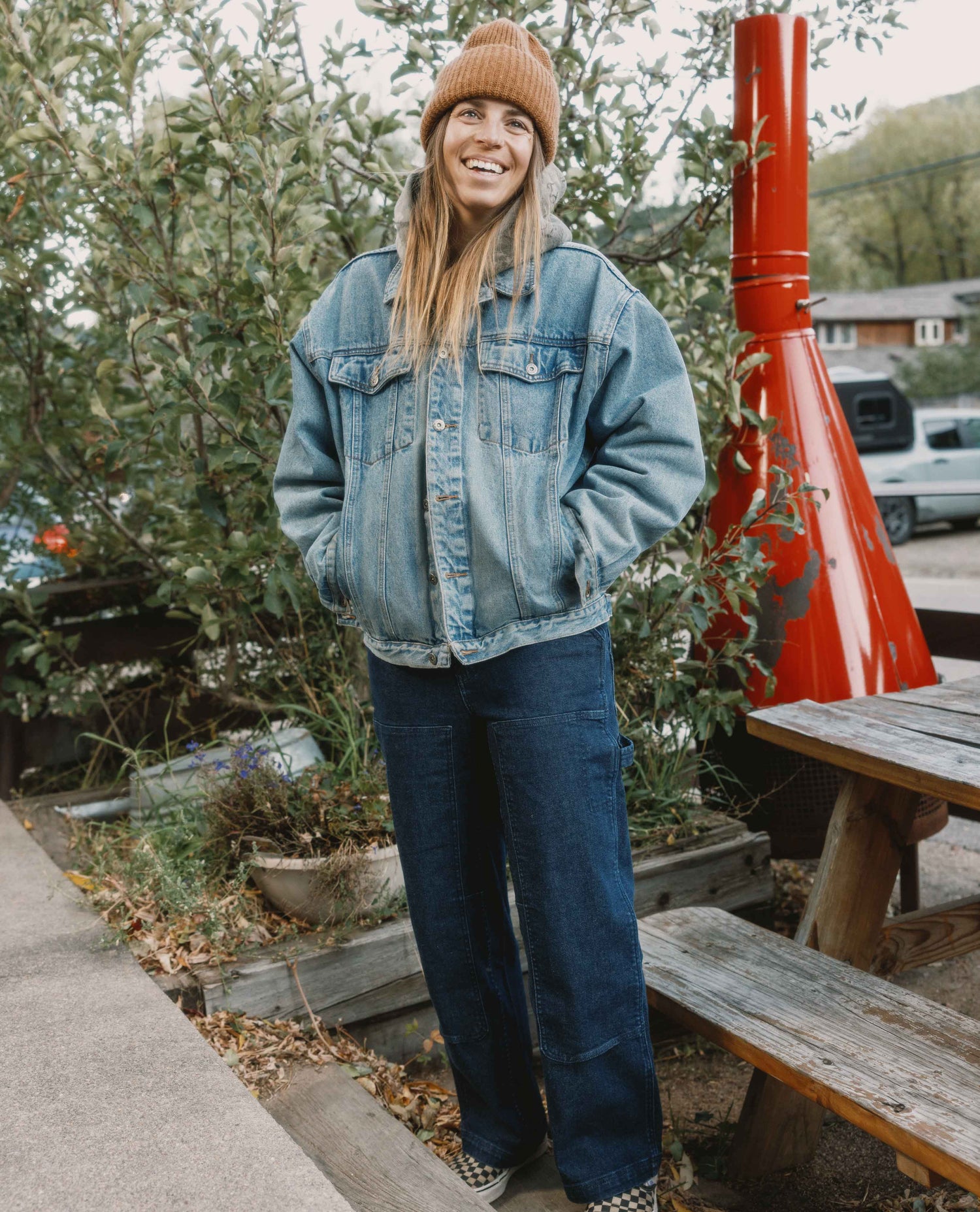 Person wearing a denim jacket and beanie standing outdoors with greenery and a red structure in the background.