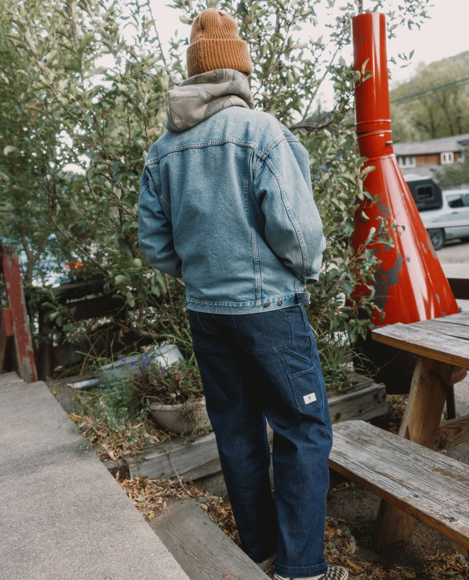 Person wearing a denim jacket and beanie standing outdoors with trees and a red structure in the background.