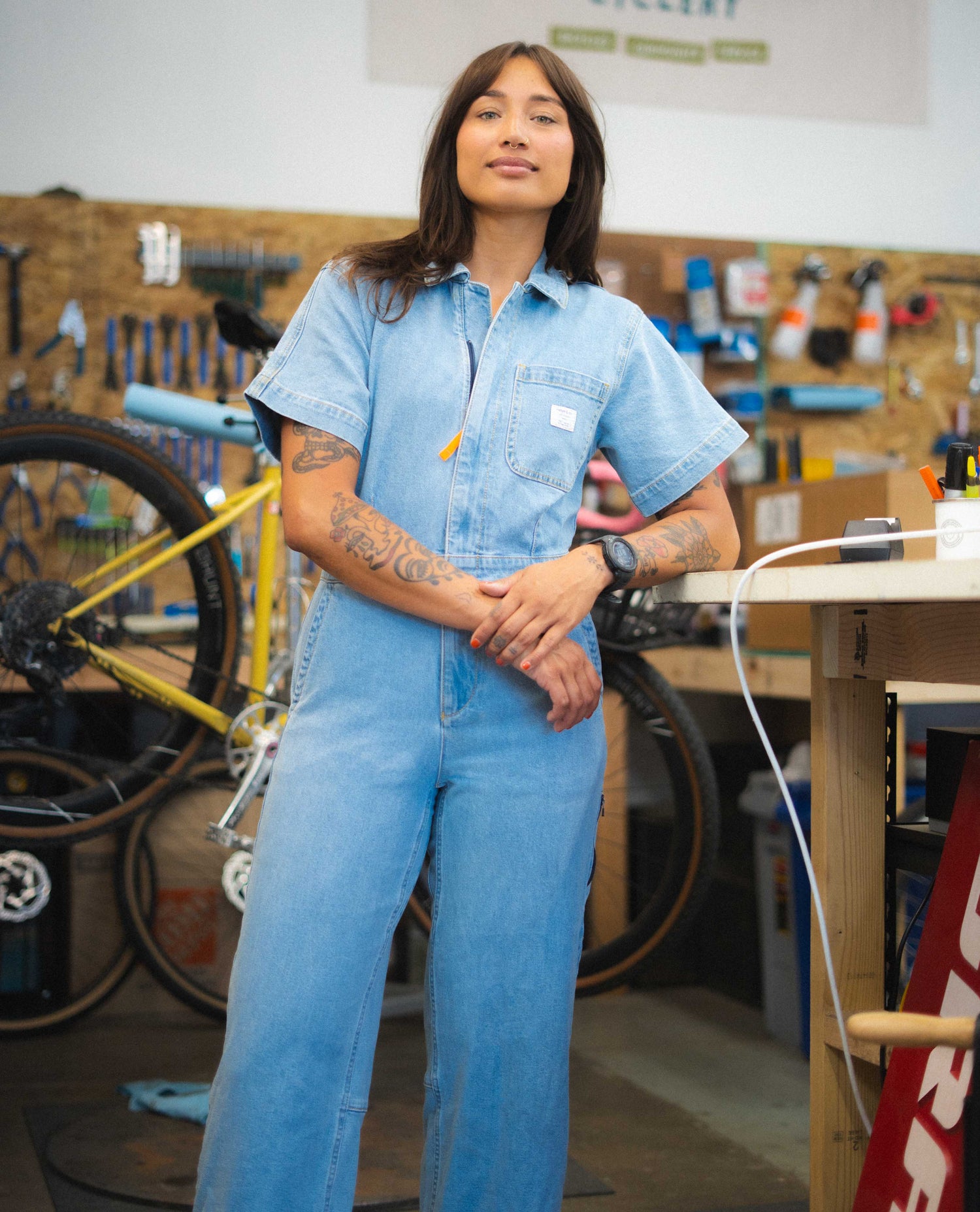 Person wearing a blue jumpsuit in a workshop setting with tools and bicycles in the background.
