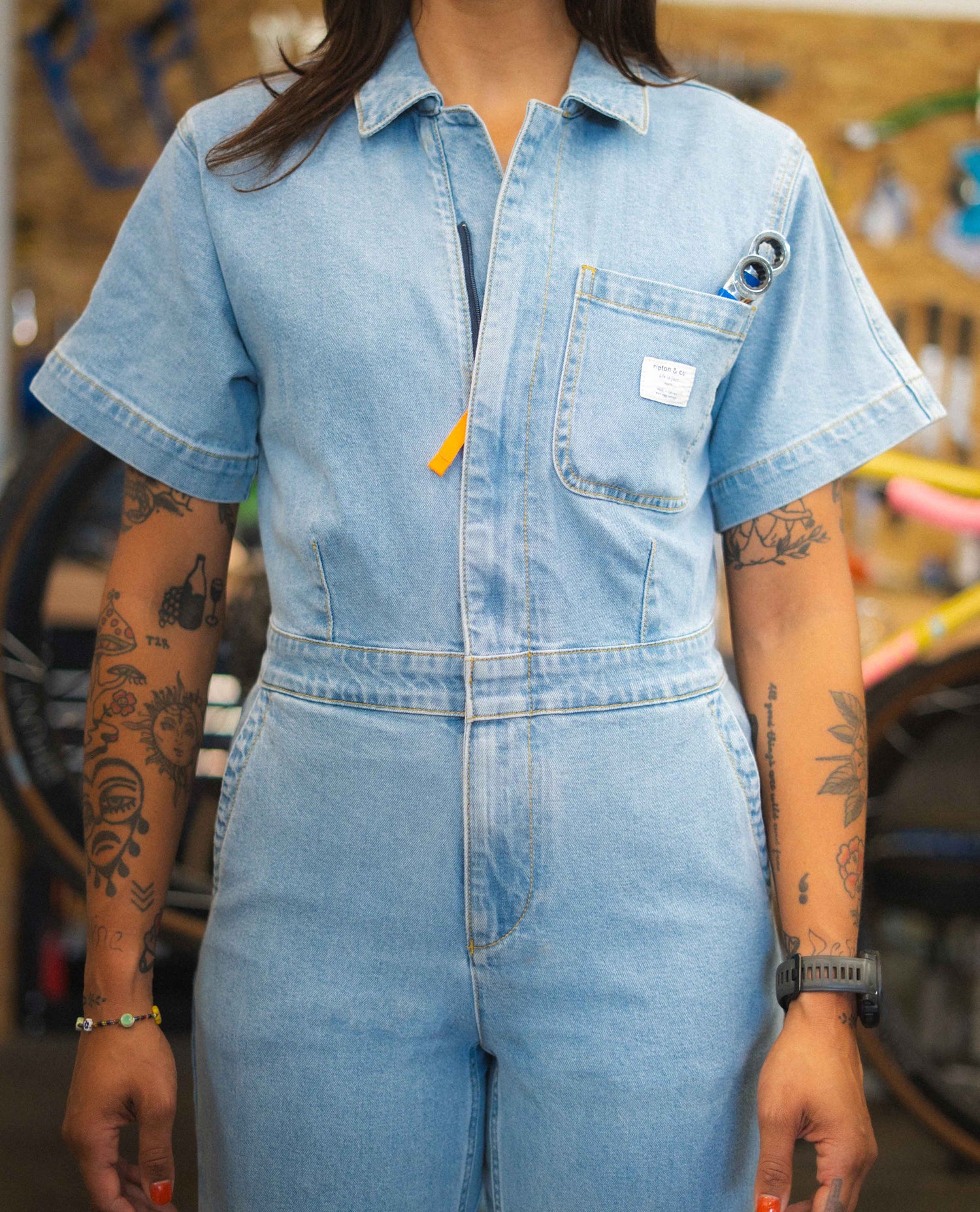 Person wearing a light blue denim jumpsuit with tattoos visible on arms, in an indoor setting.