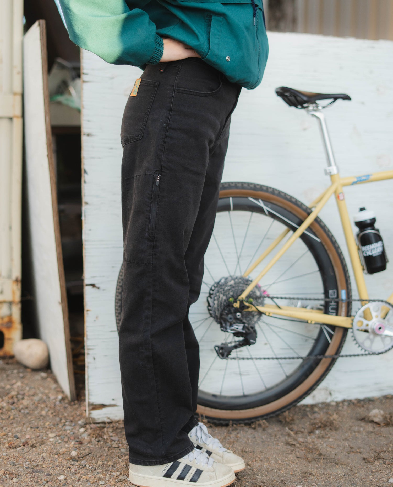 Person wearing black pants standing next to a yellow bicycle against a white wall.