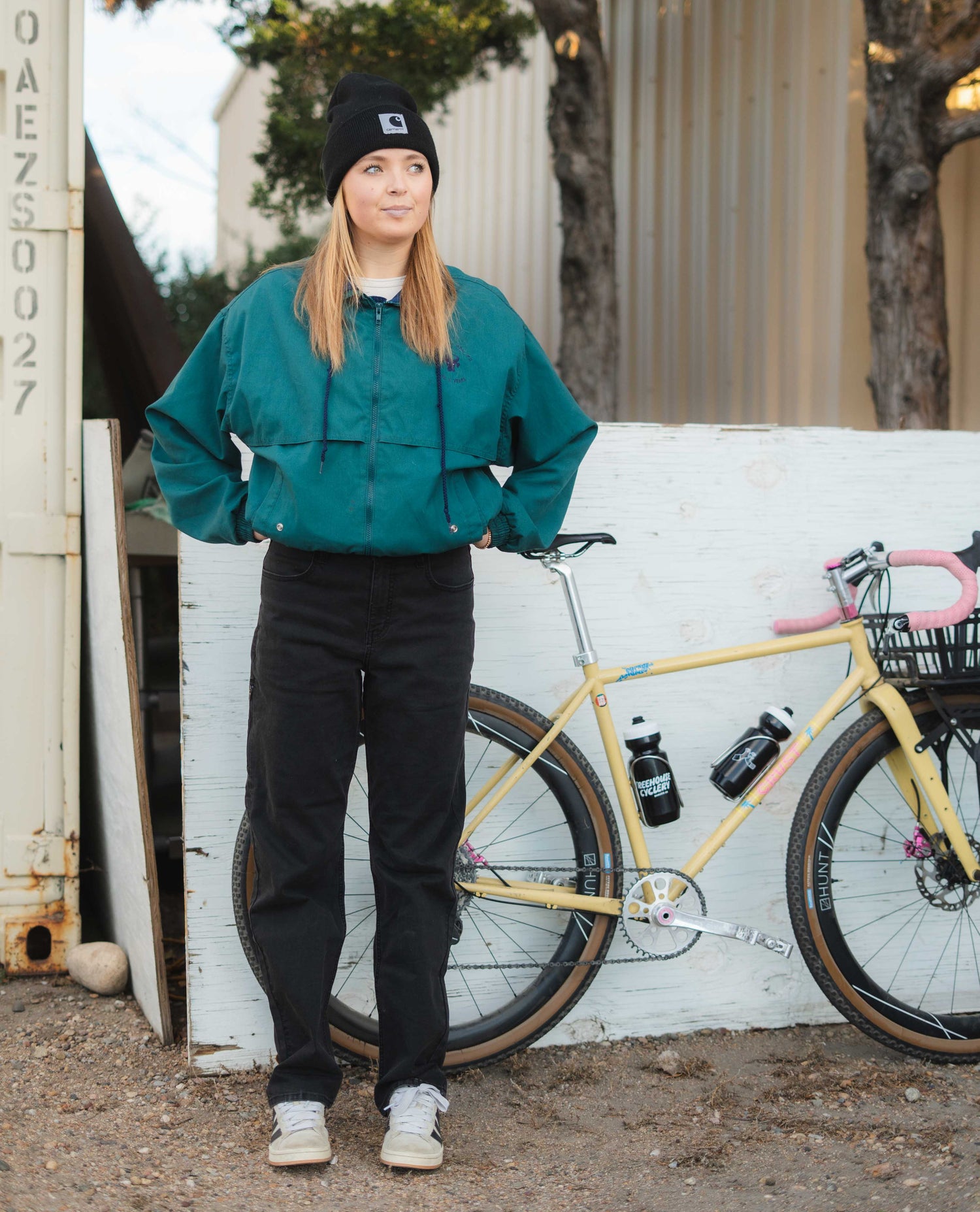 Person wearing a teal jacket and black pants standing next to a yellow bicycle with a white wall.