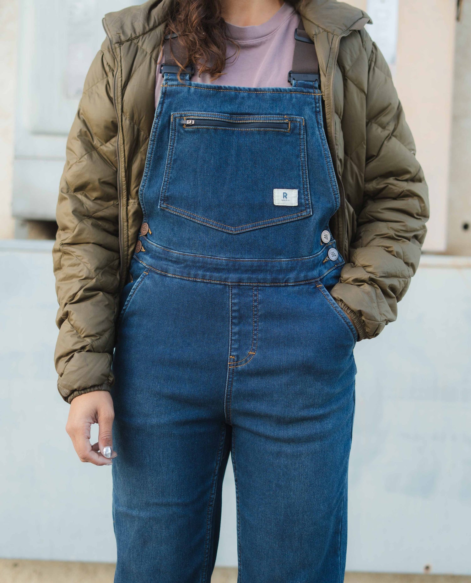 Person wearing blue denim overalls and a green jacket against a neutral background