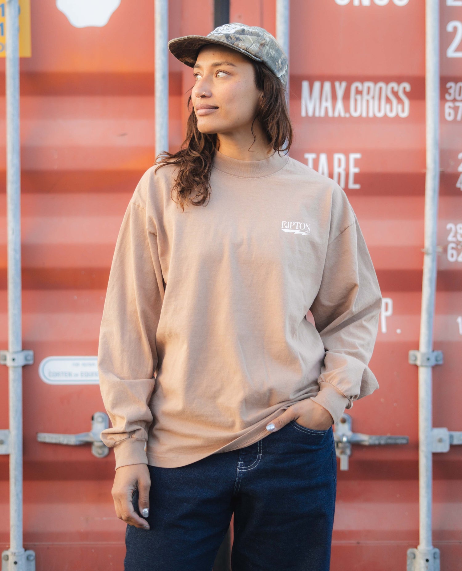 Person wearing a beige sweatshirt with a logo in front of red shipping containers.