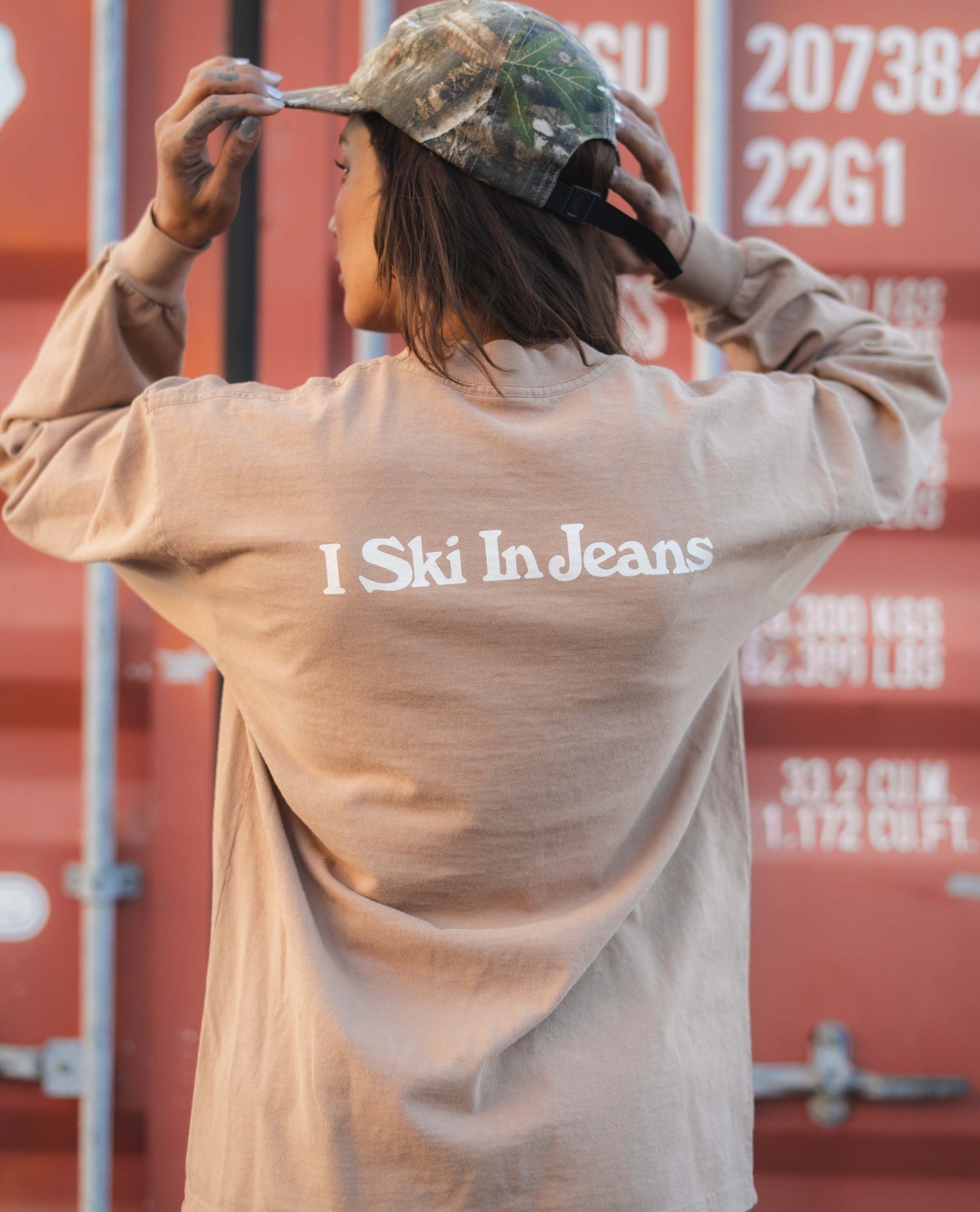 Person wearing a beige shirt with 'I Ski In Jeans' text, adjusting a cap against a red shipping container background.