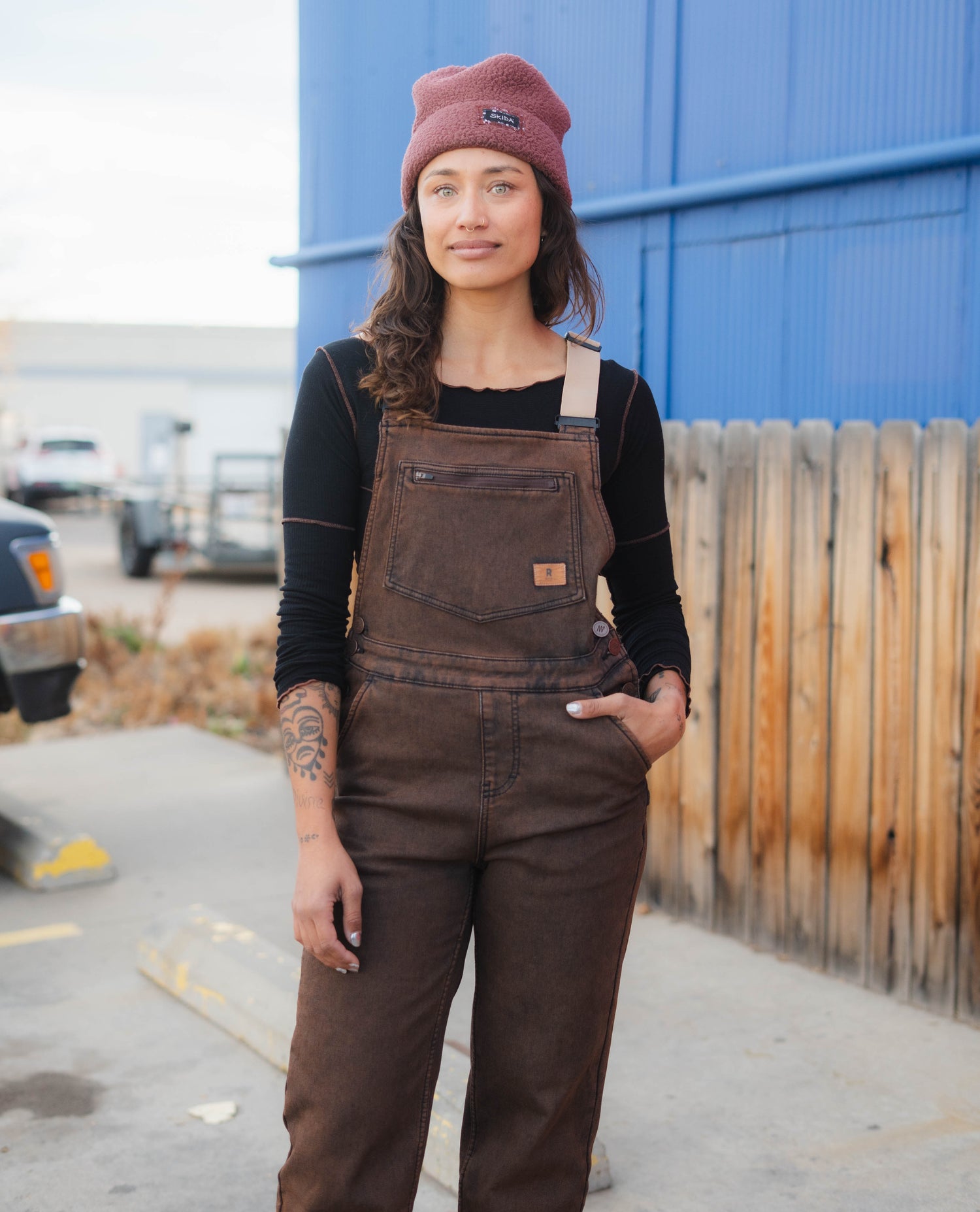 Person wearing brown overalls and a maroon beanie standing outdoors.