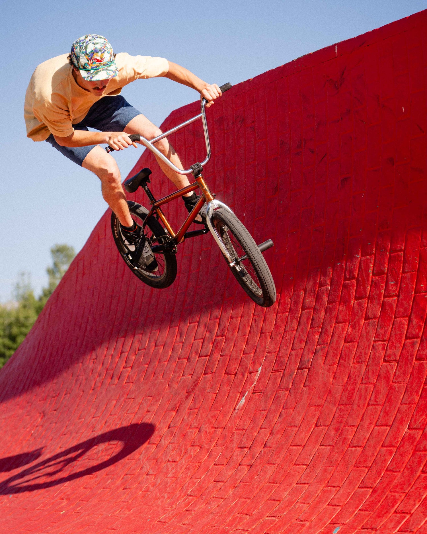 Man riding a bmx bike in a yellow shirt and Ripton jorts. He is doing a 180 on a red brick quarter pipe. 