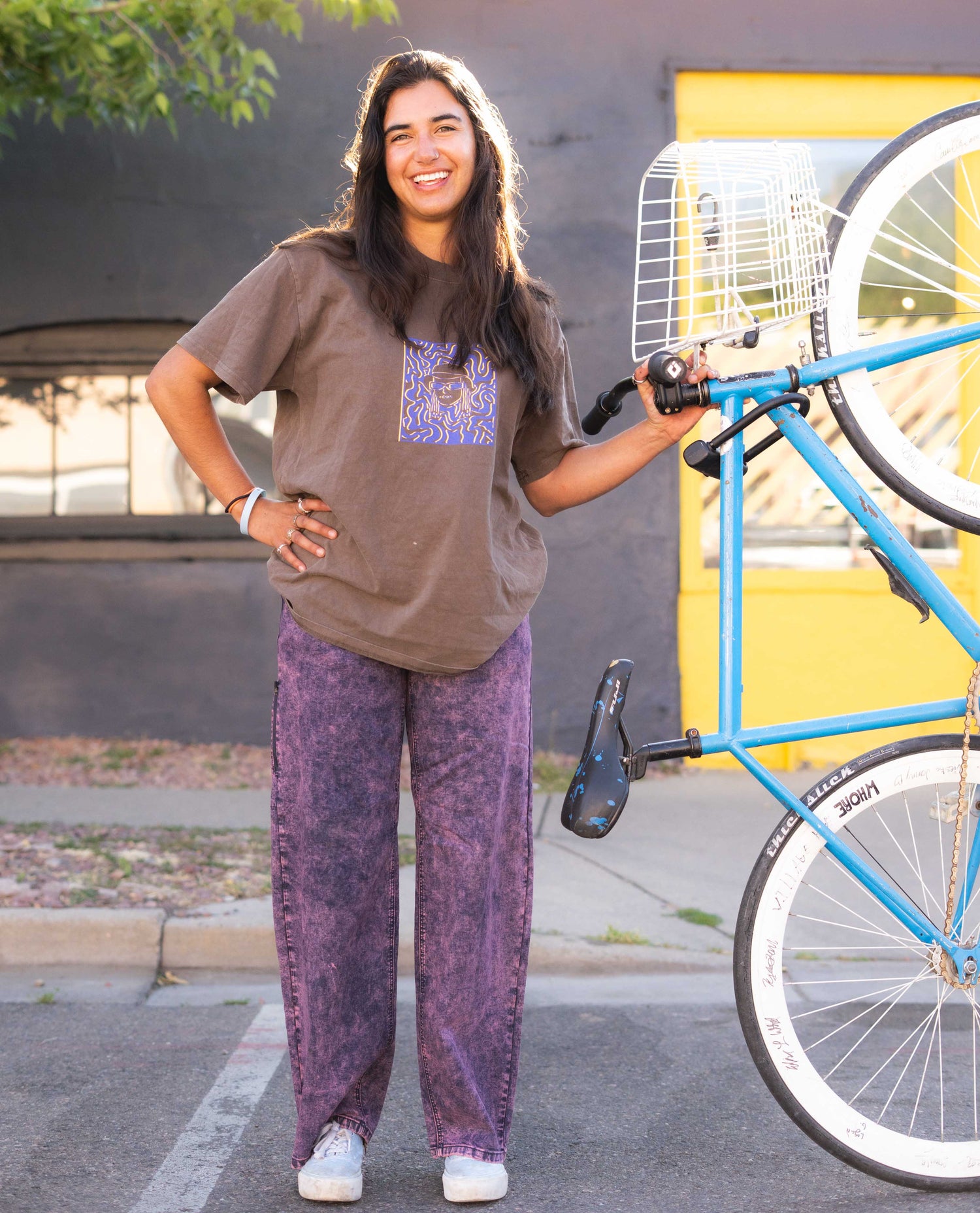 Person wearing a brown oversized t-shirt and purple pants standing next to a blue bicycle.