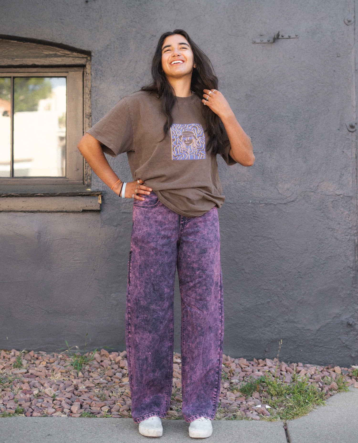 Person wearing a brown t-shirt with a graphic design and purple tie-dye pants standing against a gray wall.