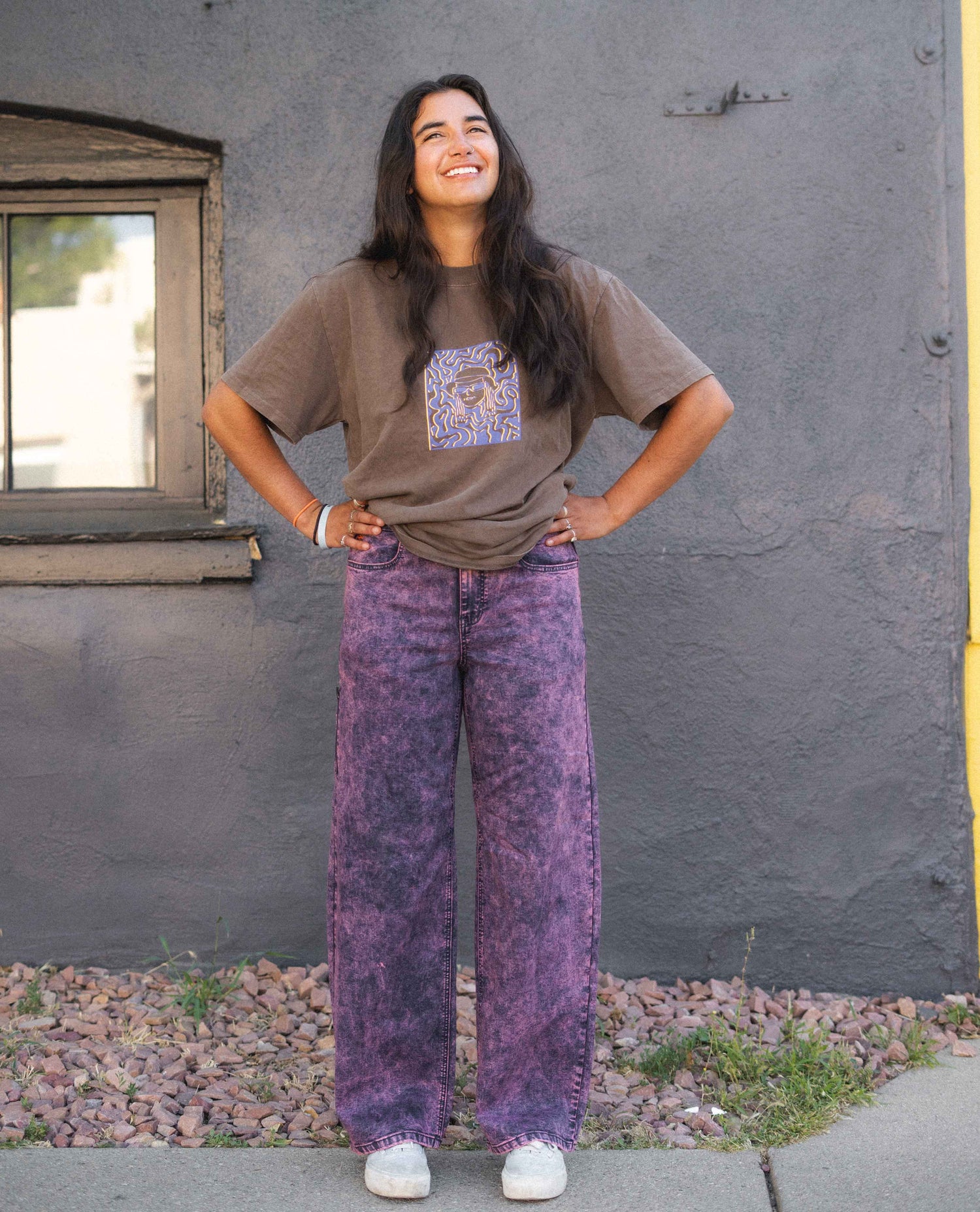 Person wearing a brown t-shirt with a graphic design and purple acid-wash jeans standing against a gray wall.