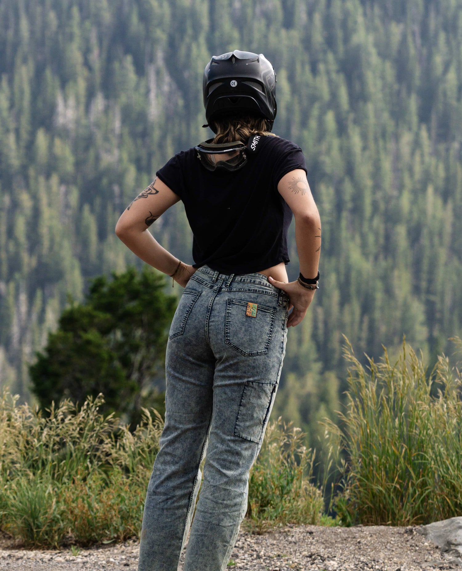 A woman wearing a black full face helmet with goggles hanging around her neck. She is also wearing a black shirt and dappled blue / grey jeans. 