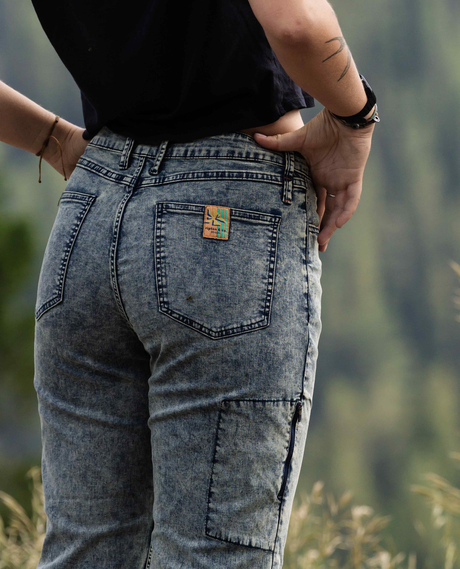 A person models a dappled blue / grey jean with a tucked in black t-shirt. Her arms show a small tattoo. 