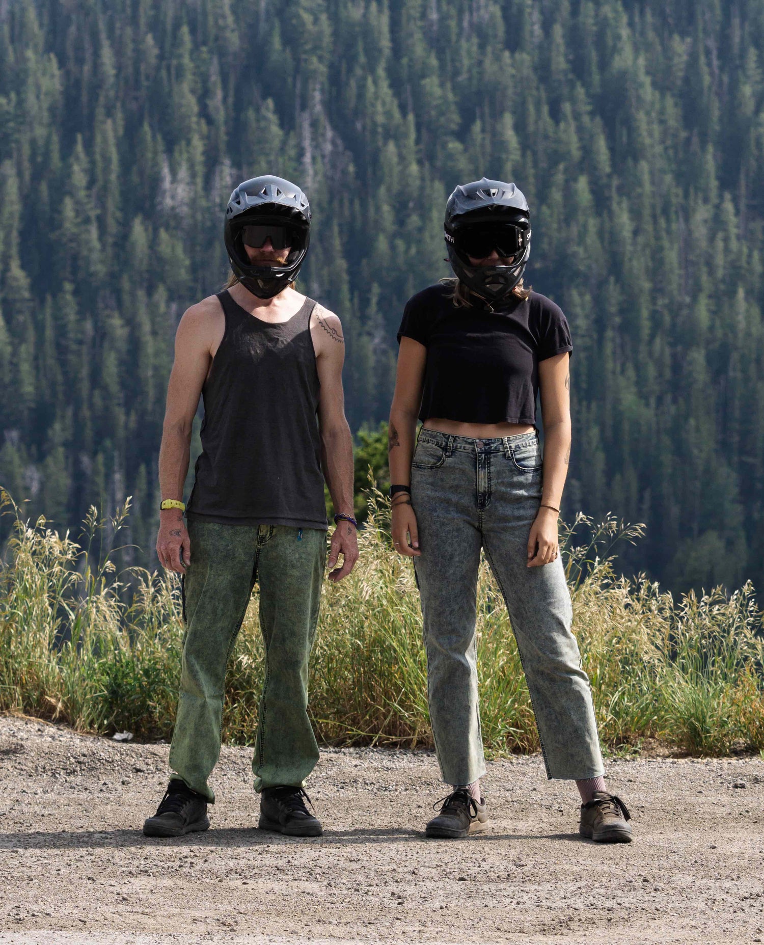 A man (left) and woman (right) in black mountain bike helmets, and black shirts looking at camera in front of a forest. Person on the left is wearing green jeans and the person on the right is wearing blue jeans. 