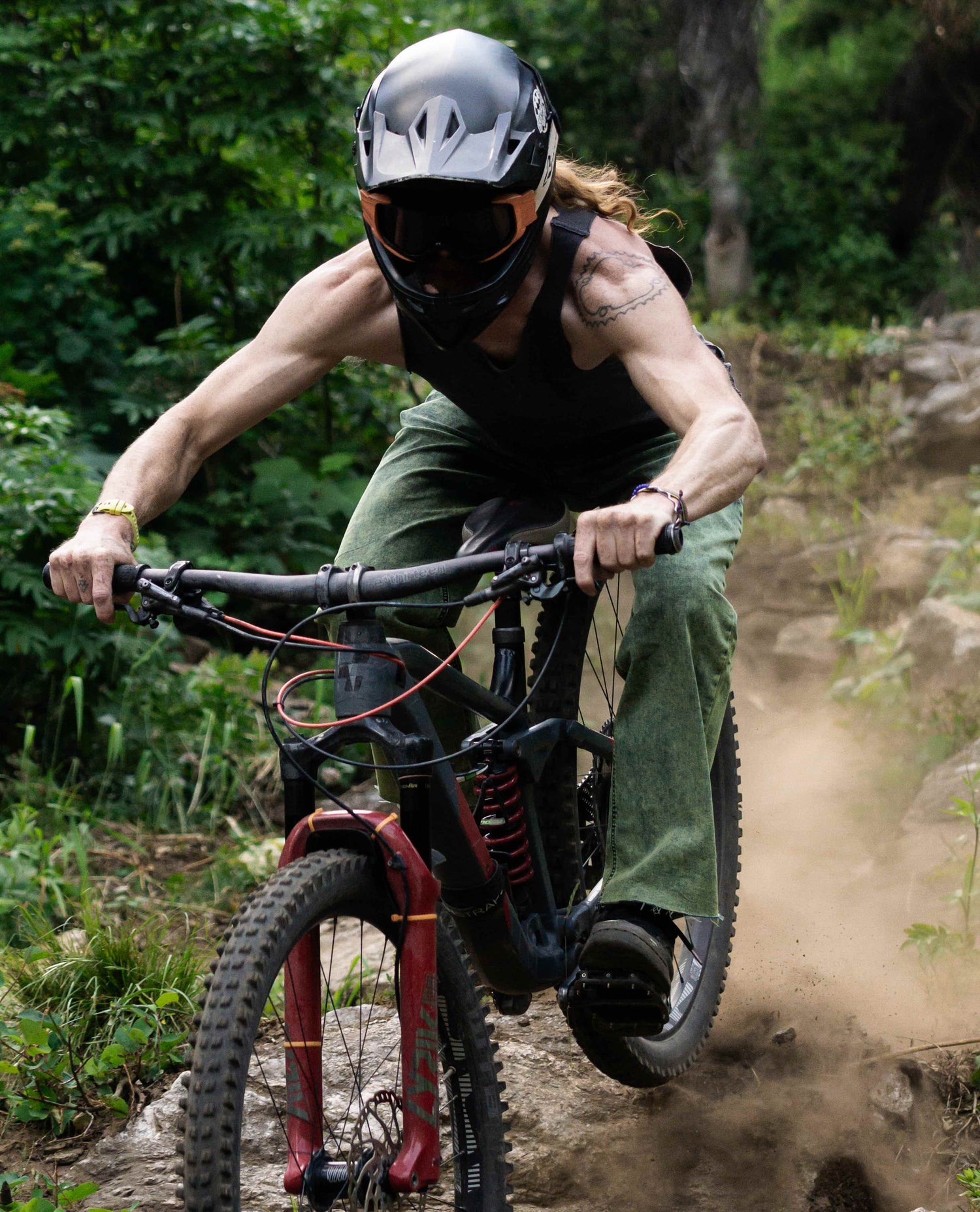 A man in a black helmet, black tank top and green jeans riding his mountain bike over rocks in the woods. Dust sprays from behind his rear tire. 