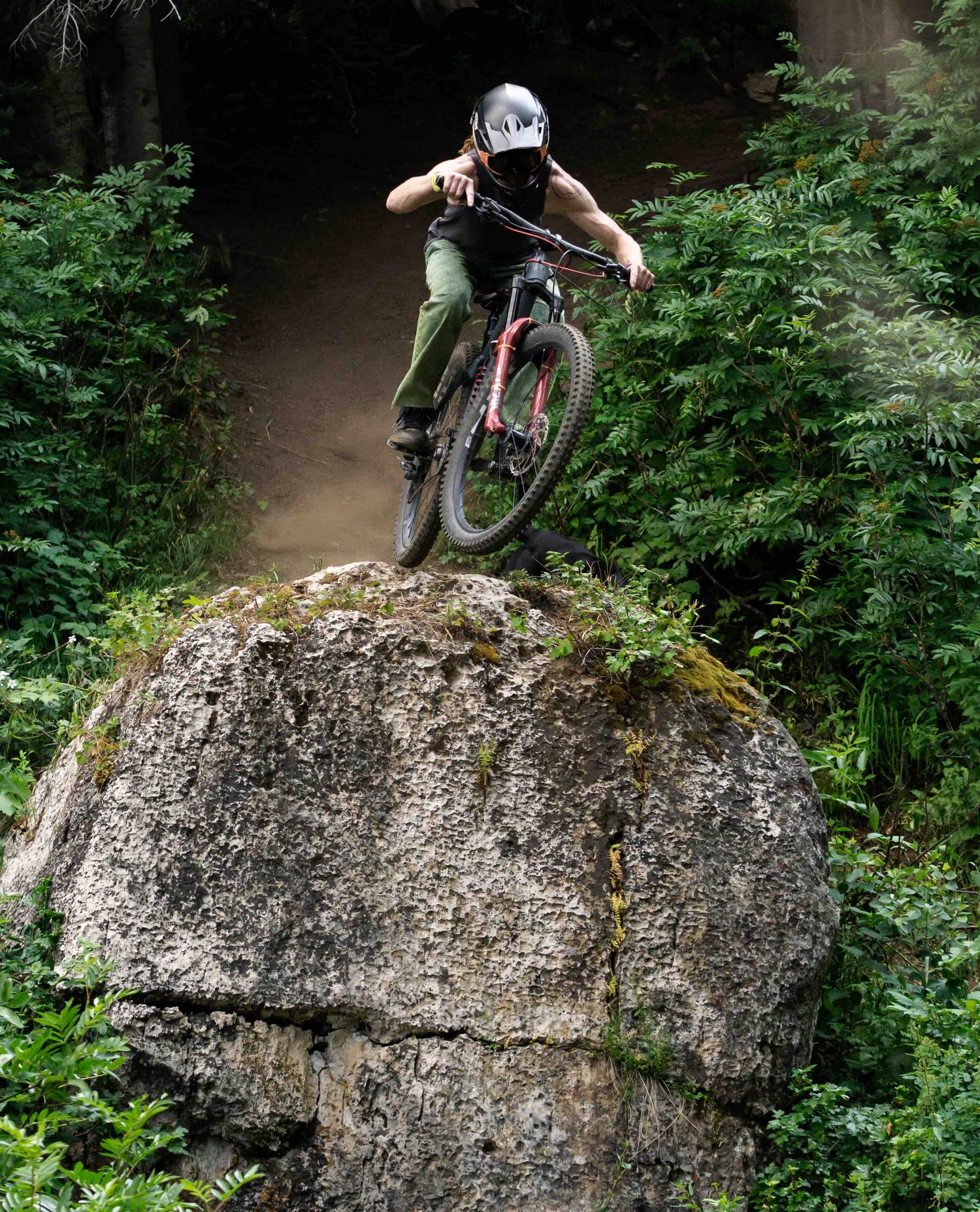 A man in a black helmet, black tank top and green jeans riding his mountain bike off of a large boulder in the woods.