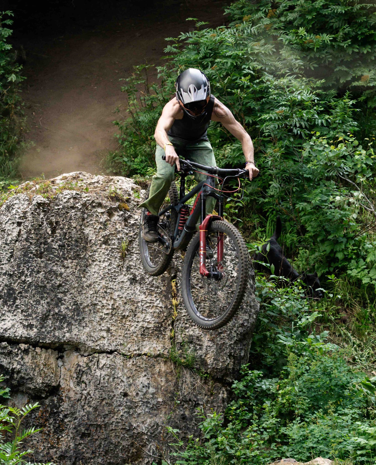 A man in a black helmet, black tank top and green jeans riding his mountain bike off of a large boulder in the woods.