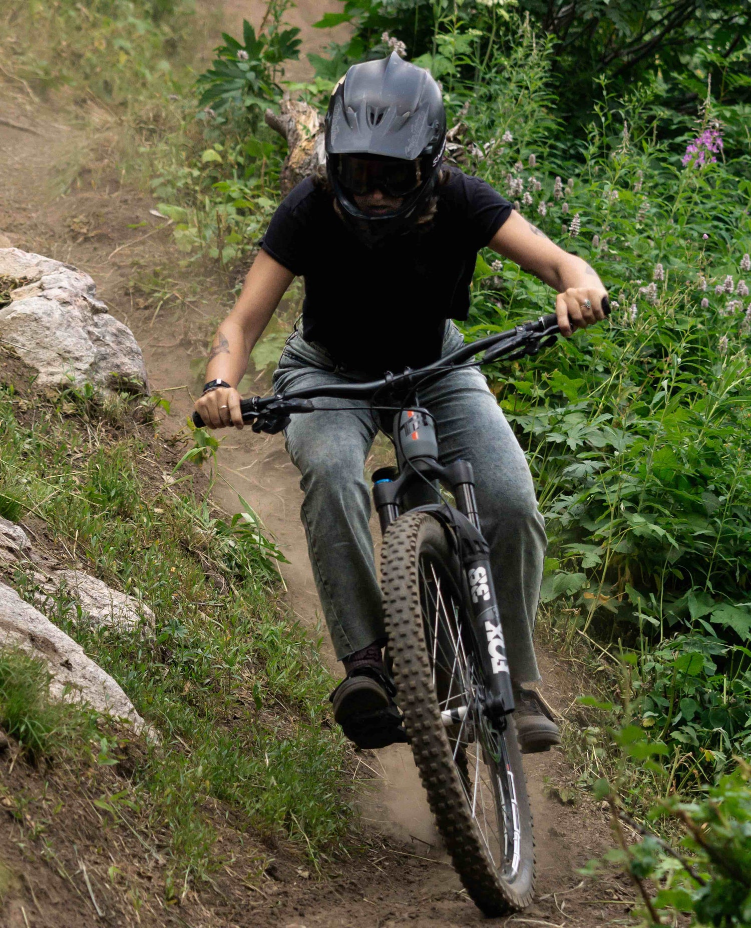 A female mountain biker in a black full face helmet, black t-shirt, and light grey jeans rides her mountain bike in the woods. Dust trails her. 