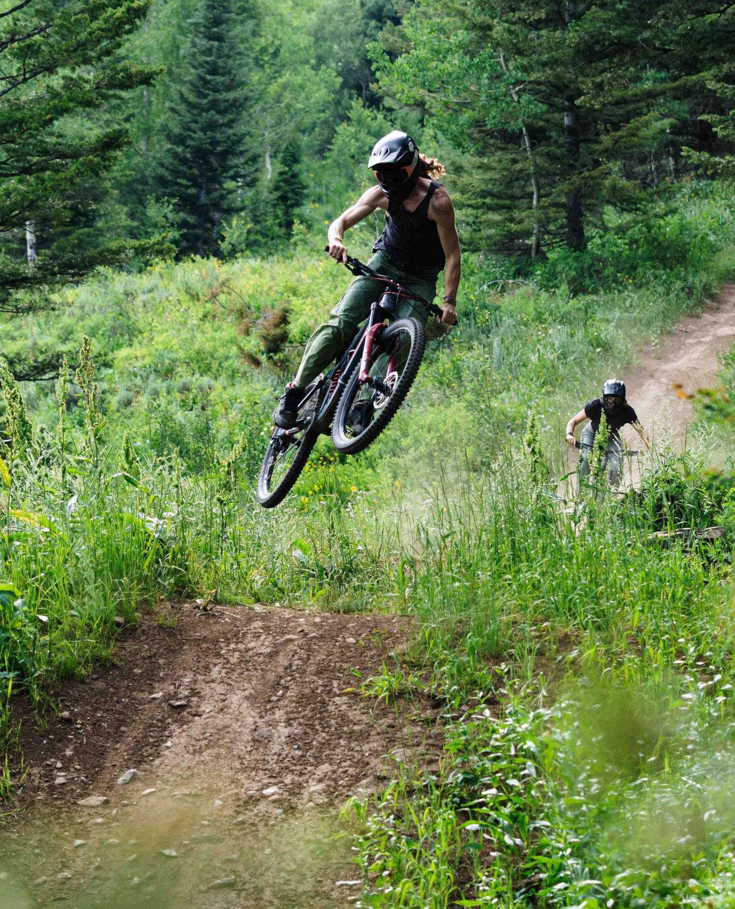 A man wearing a black full face helmet, black tank top, and green jeans jumping his mountain bike in the woods. He is followed by a female rider also wearing a black full face helmet and a black t-shirt. 