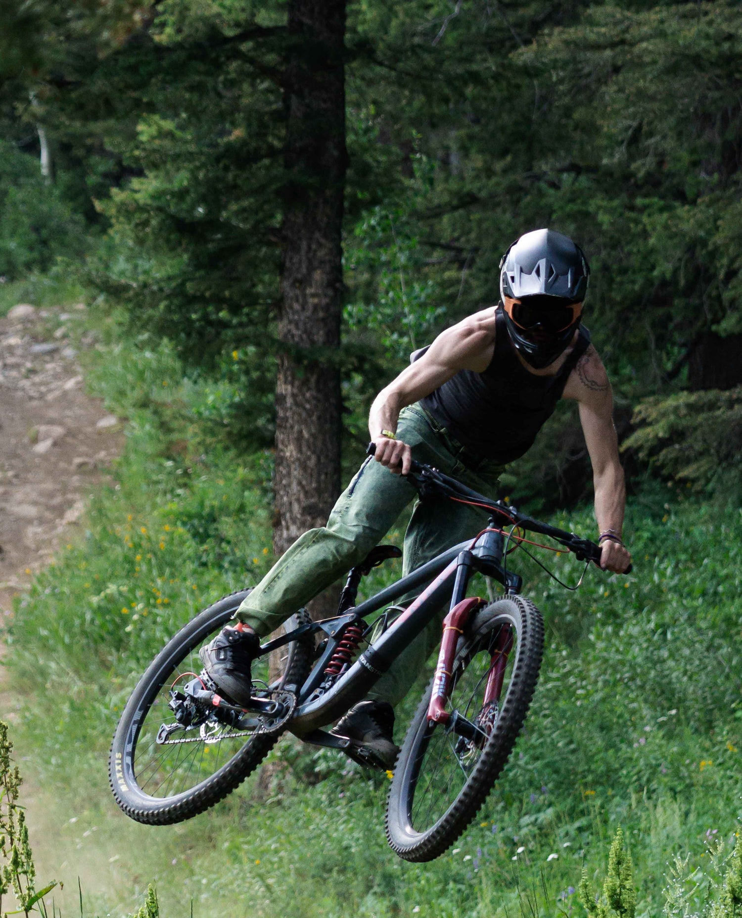 A man wearing a black full face helmet, black tank top and green jeans jumping his mountain bike in the woods. 