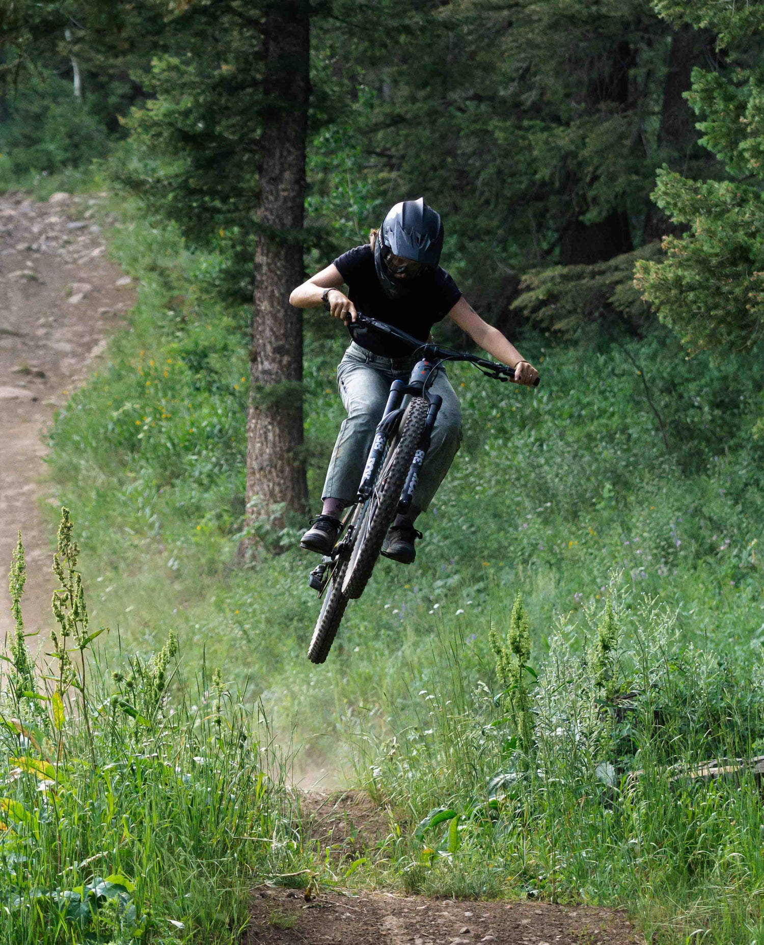 A female mountain biker wearing a black full face helmet, black t-shirt, and blue jeans jumps her mountain bike in the woods. 