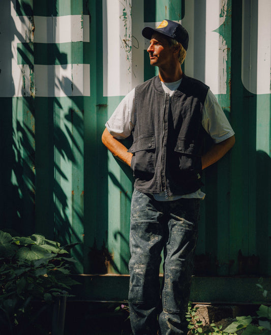 A man wearing a dark gray utility vest over a white t-shirt, blue jeans, and a dark baseball cap stands in front of a weathered green and white corrugated metal wall with dappled sunlight and shadows.