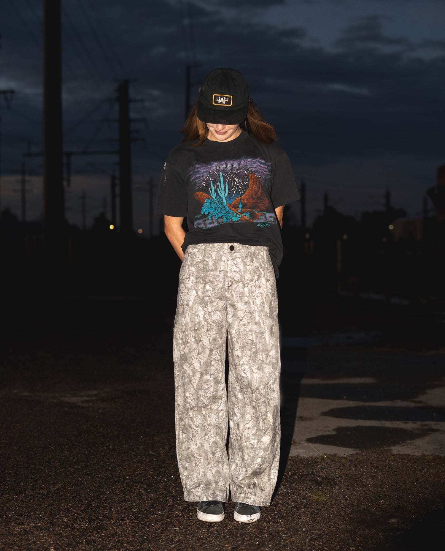 Person wearing a black t-shirt with a graphic design and patterned pants standing outdoors at night.