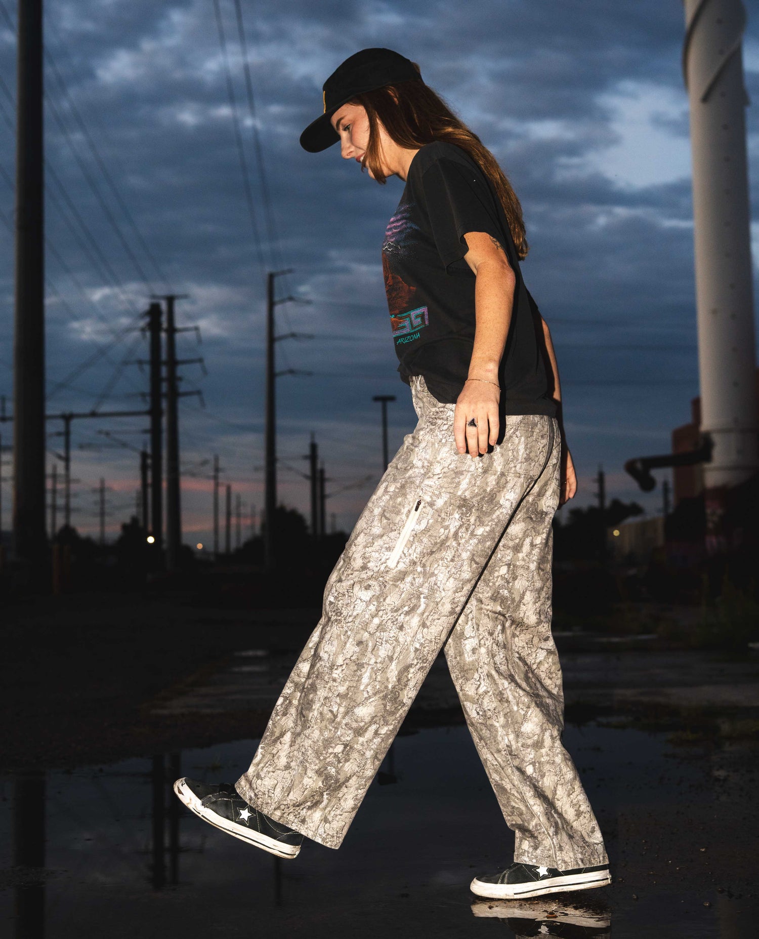 Person wearing a black cap, black t-shirt, and patterned pants walking on a street at dusk.