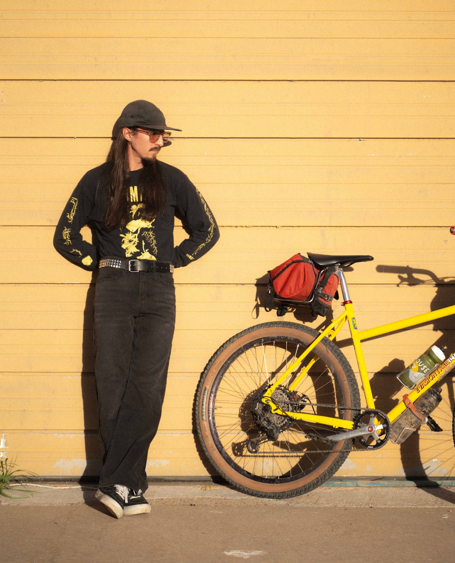 Person standing next to a yellow bicycle with a red bag against a yellow wall.