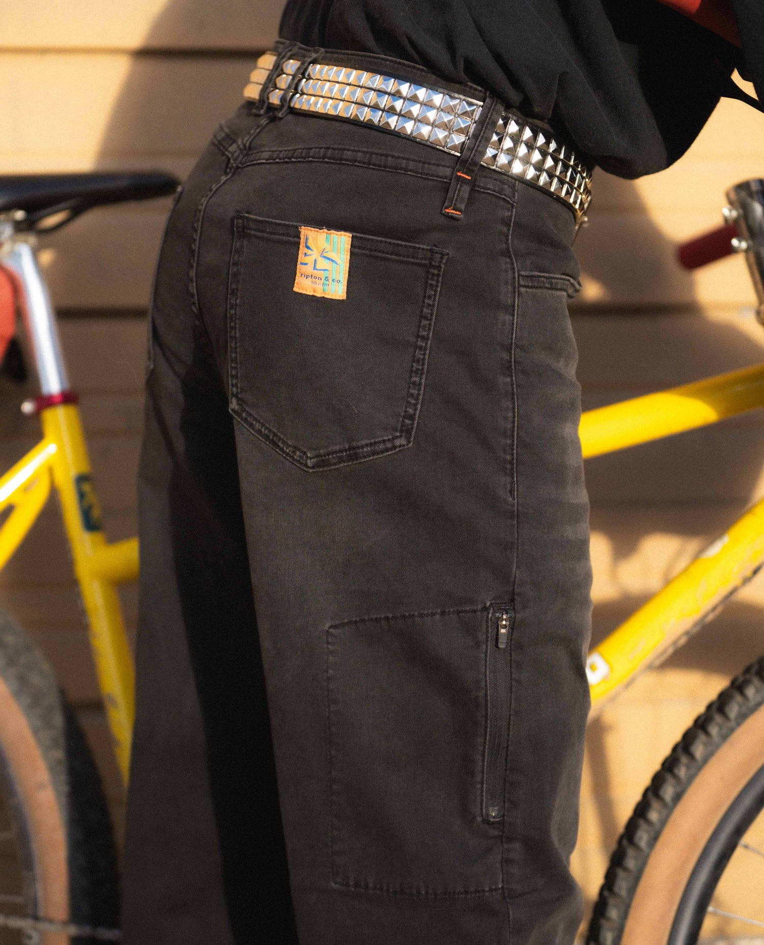 Black pants with a brand label and studded belt, yellow bicycle in the background