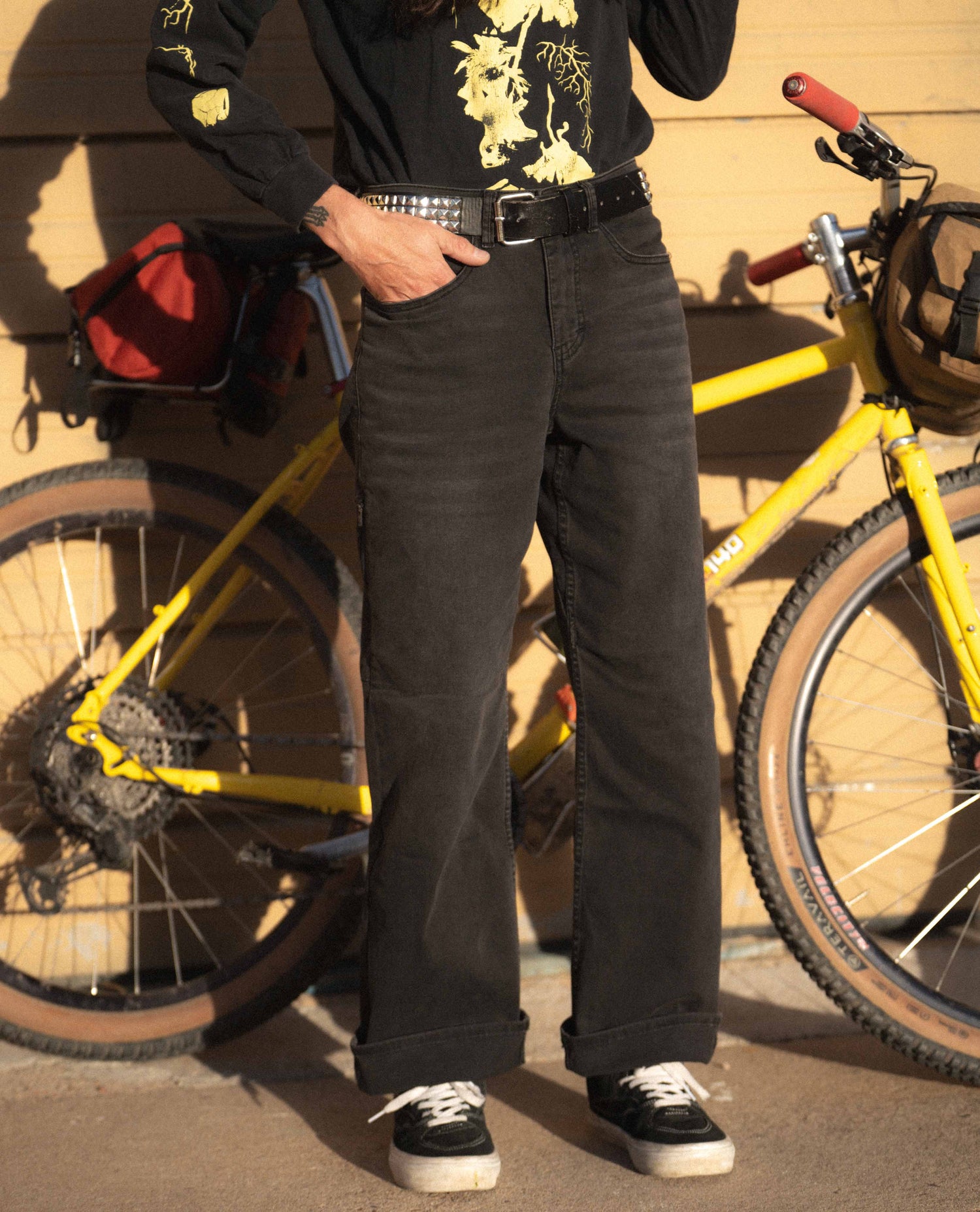 Person wearing black pants standing next to a yellow bicycle with a wooden wall background