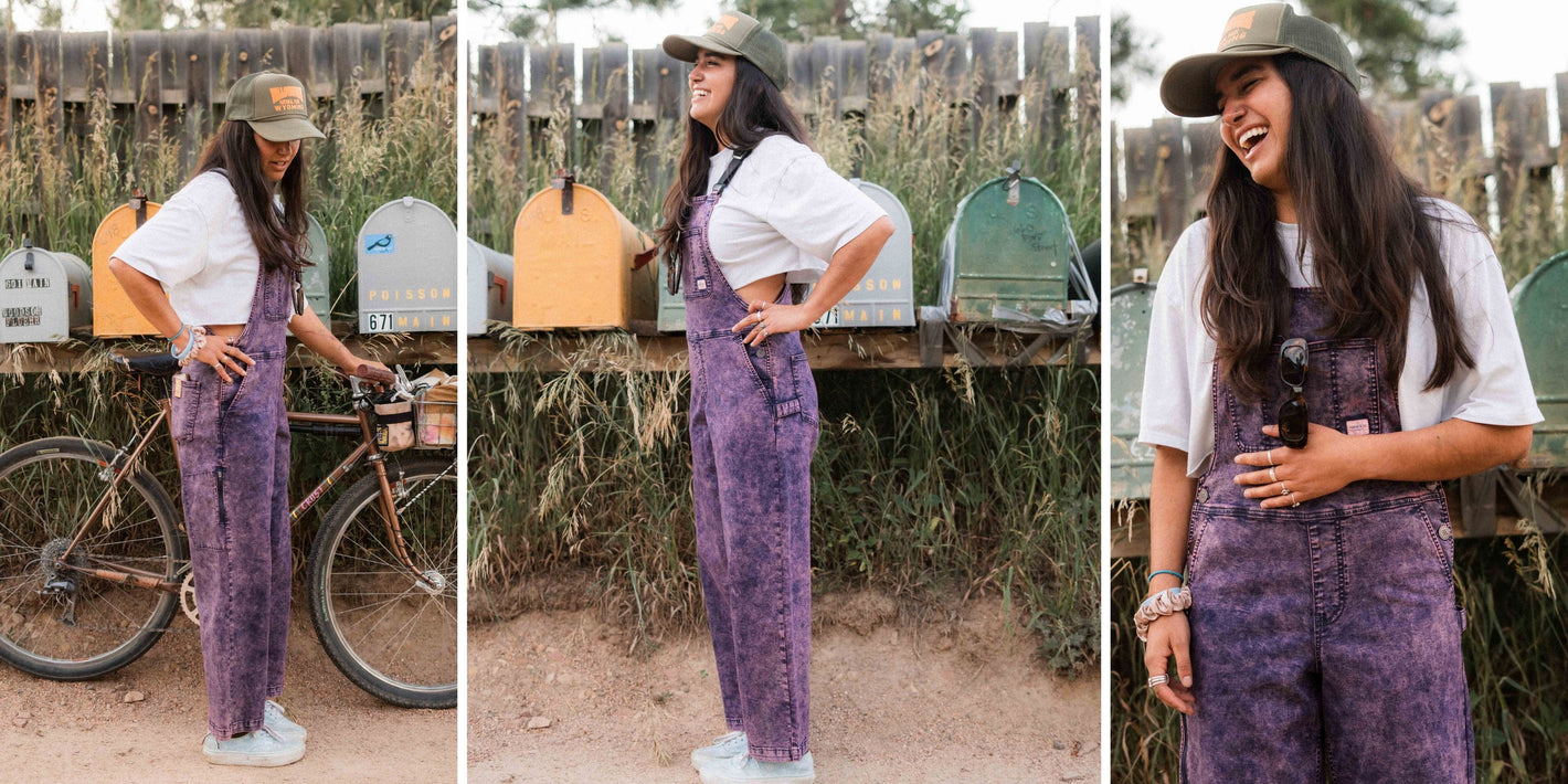 Woman in a purple denim overalls standing outdoors with a bicycle and mailboxes in the background.