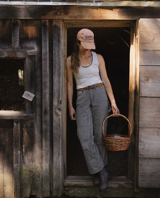 Woman stands in chicken coop entry wearing white shirt, orange hat, and black and white striped pants. She's holding a basket. 