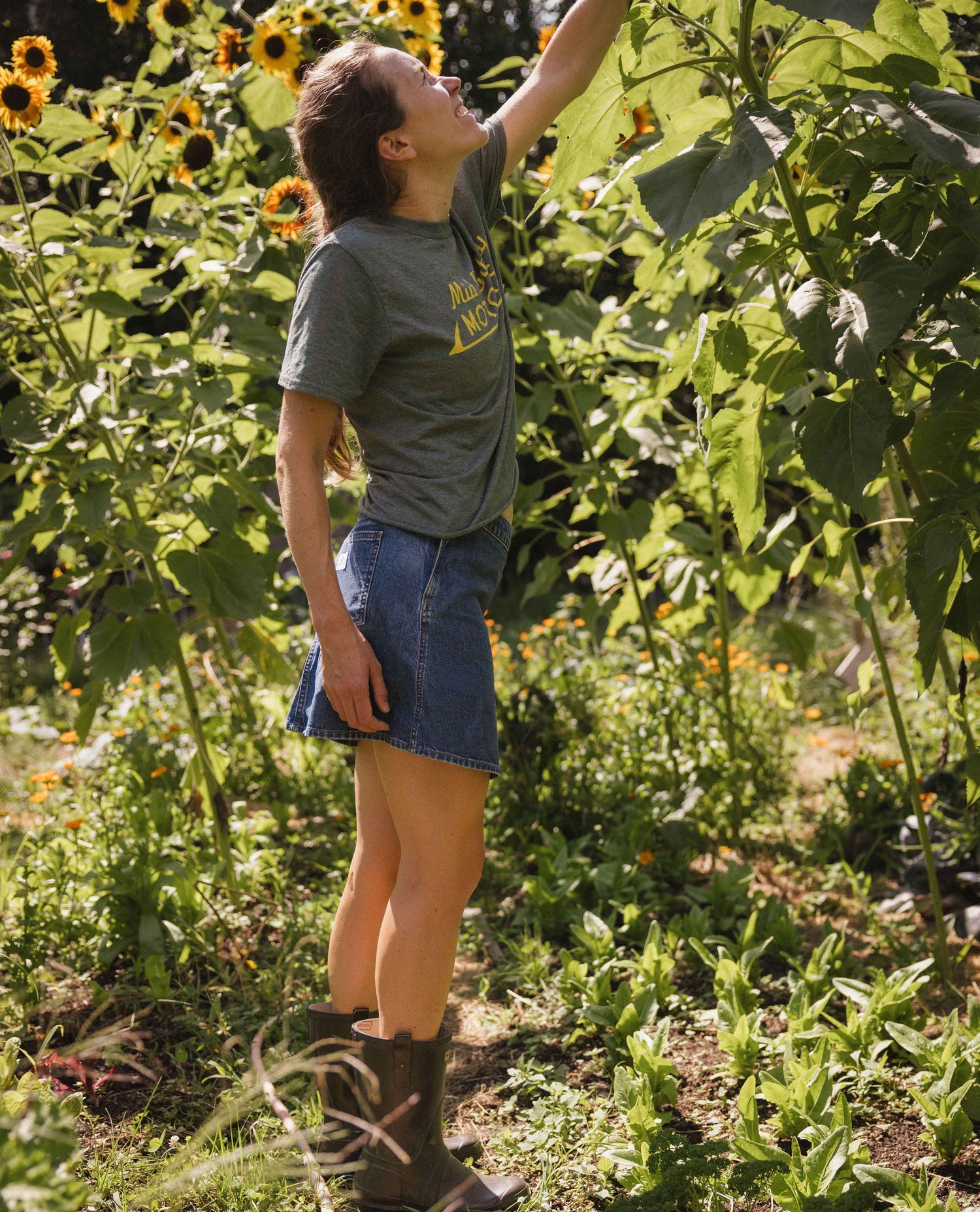 A woman wearing a gray graphic t-shirt, denim skirt, and brown boots stands in a sunflower field, reaching up toward a large sunflower plant.