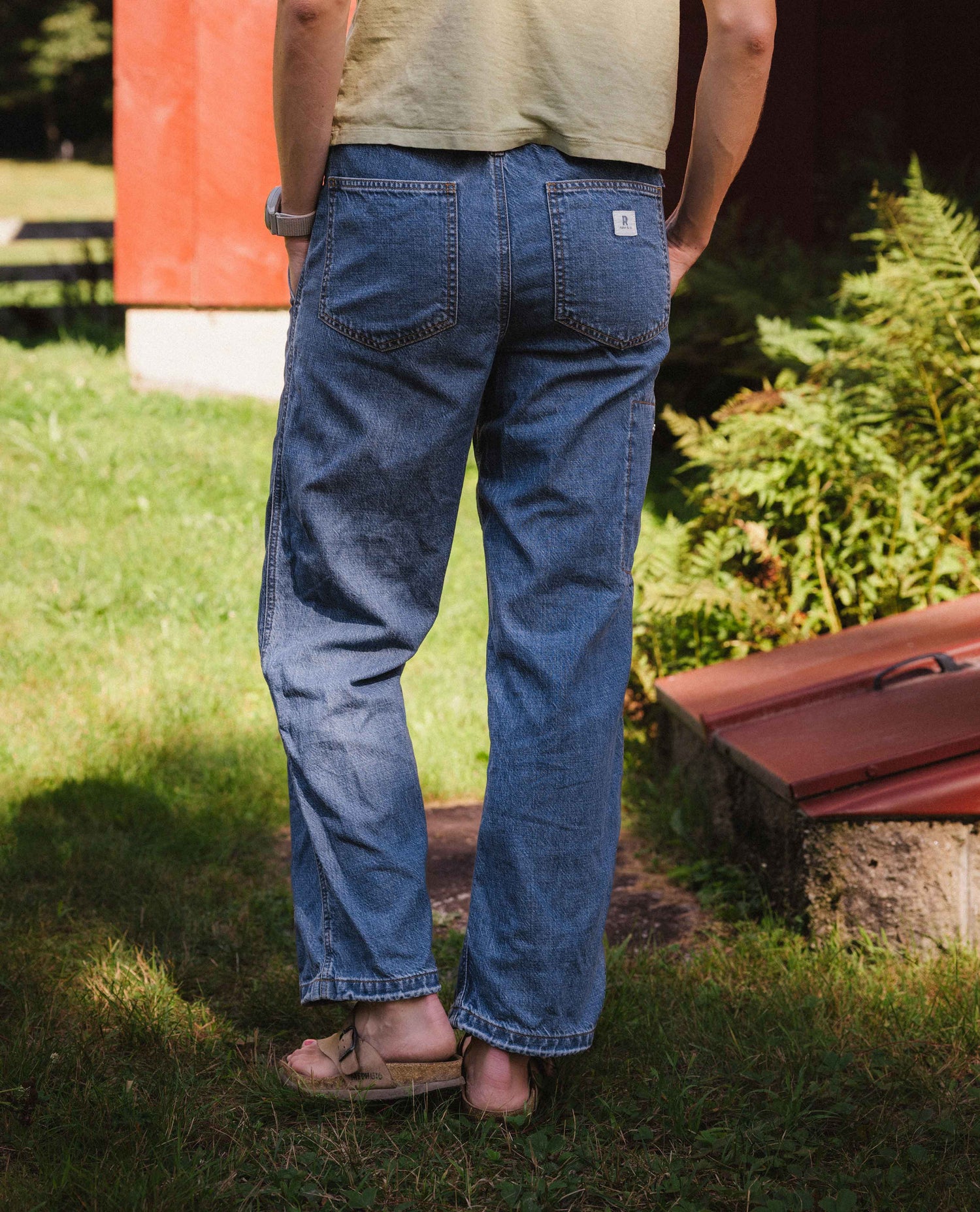 A person standing outdoors on grass, shown from the back wearing blue denim jeans with a light beige t-shirt and sandals. The jeans have a classic straight-leg fit and a small white brand patch on the upper right back pocket.