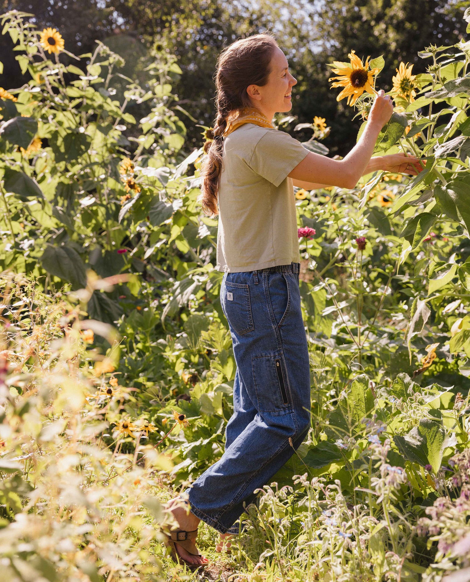 A woman stands in a sunflower field, wearing a beige short-sleeve t-shirt and blue cargo-style jeans, reaching toward a sunflower.