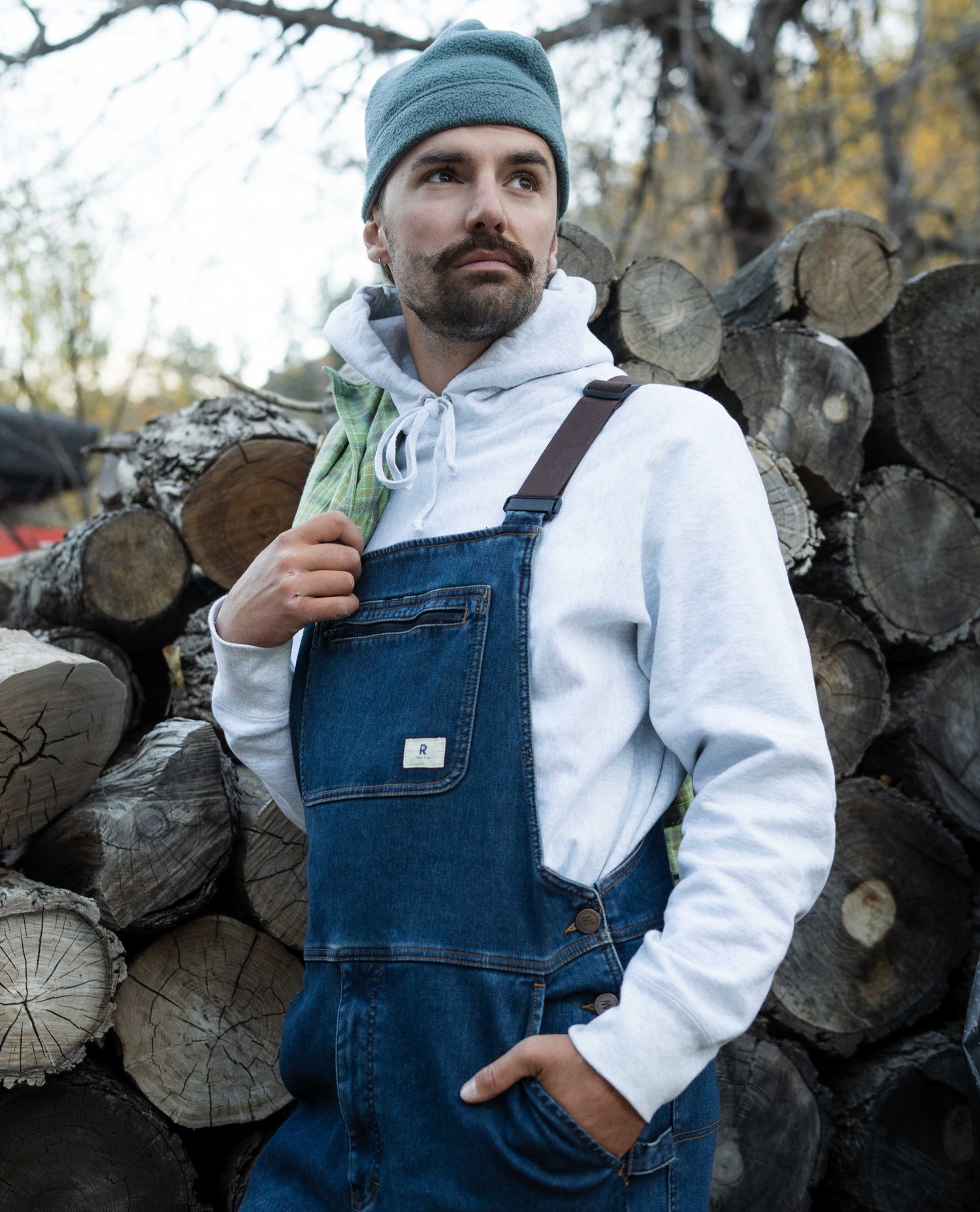 Man wearing a white hoodie, blue denim overalls, and a green beanie standing in front of stacked logs.
