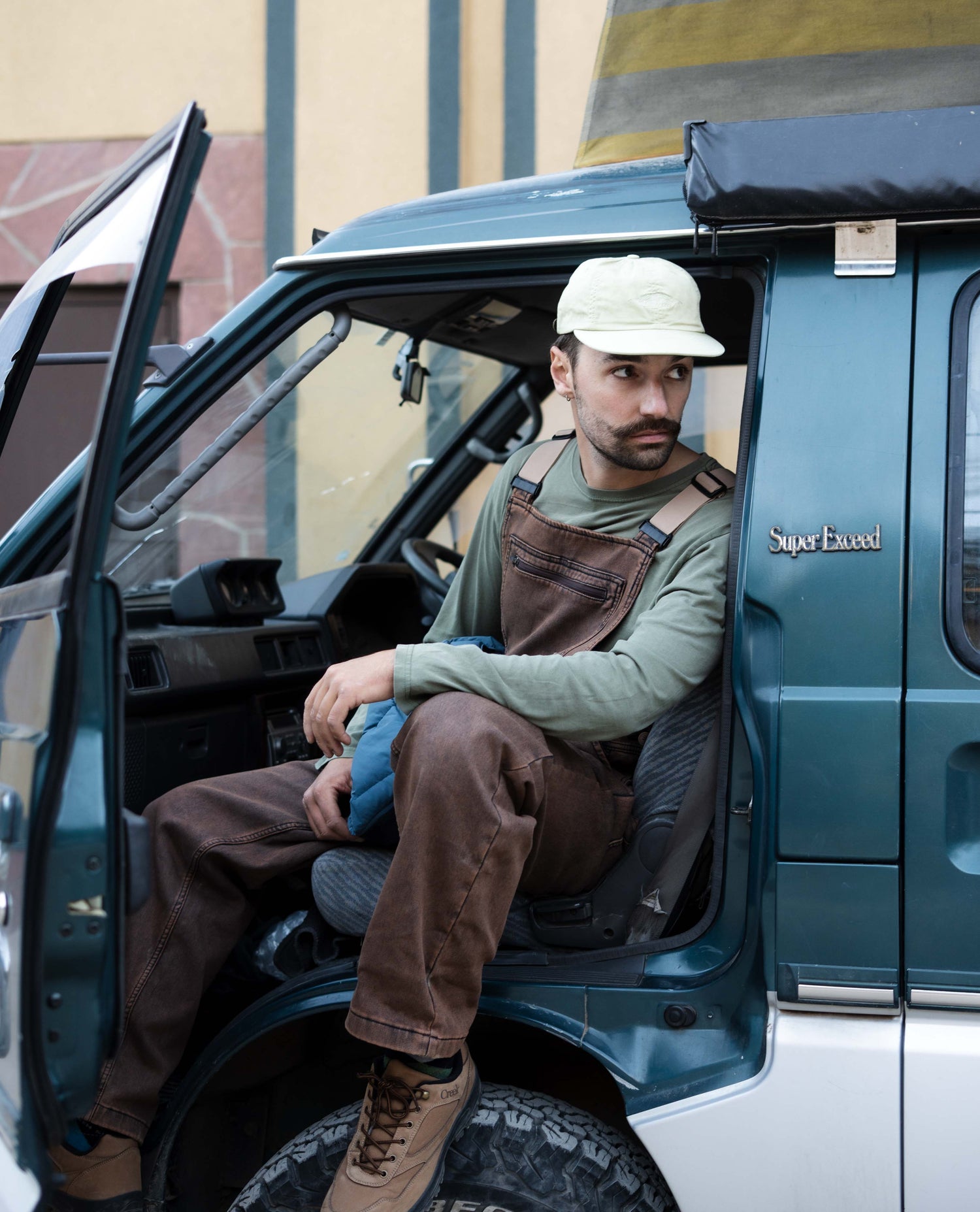 Man sitting inside a teal truck wearing brown overalls and a white cap.
