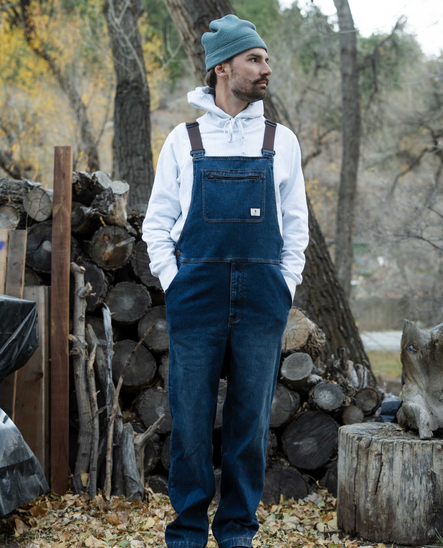 Man wearing blue overalls and a beanie standing in front of stacked logs.