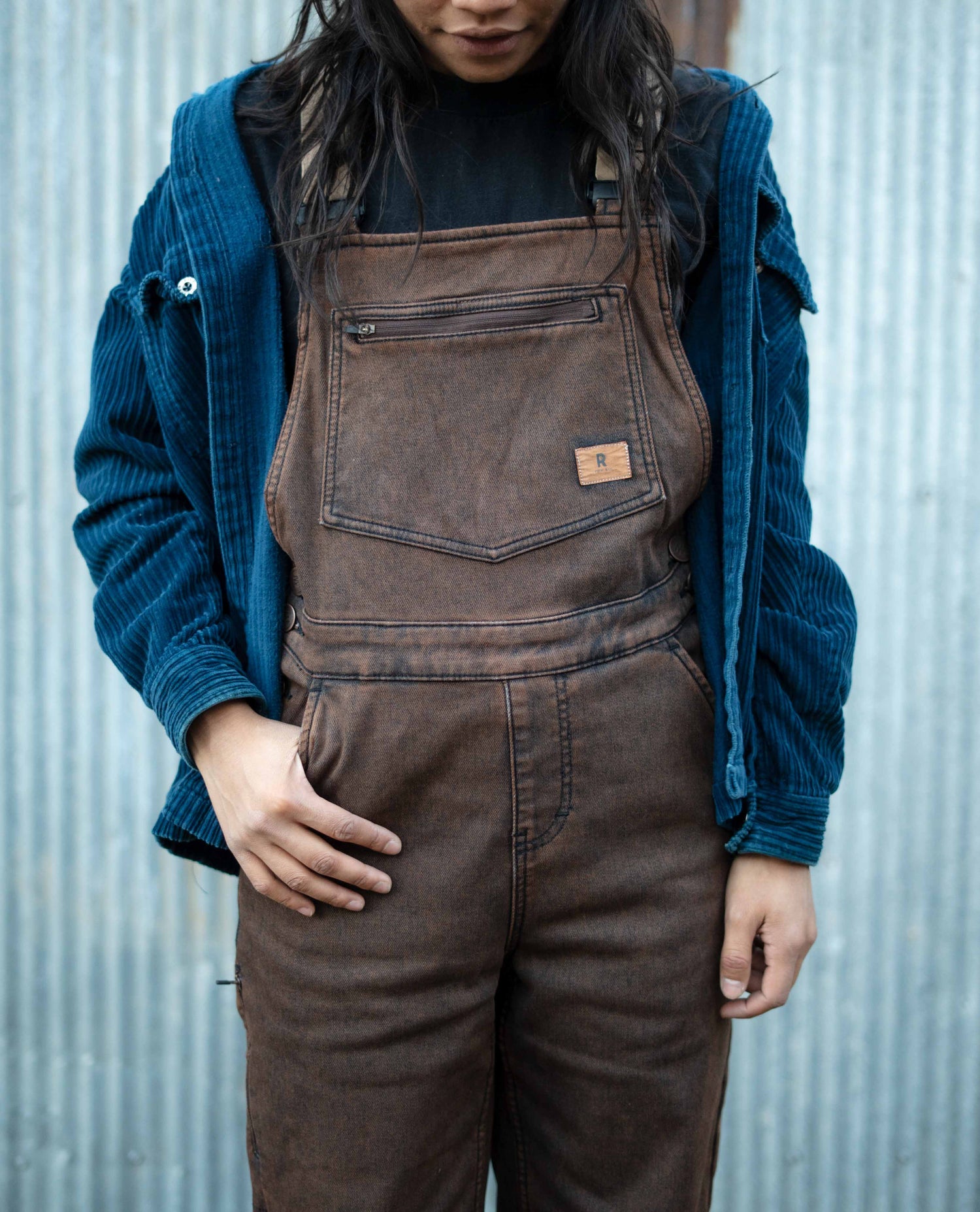 Person wearing brown overalls with a blue jacket against a corrugated metal background