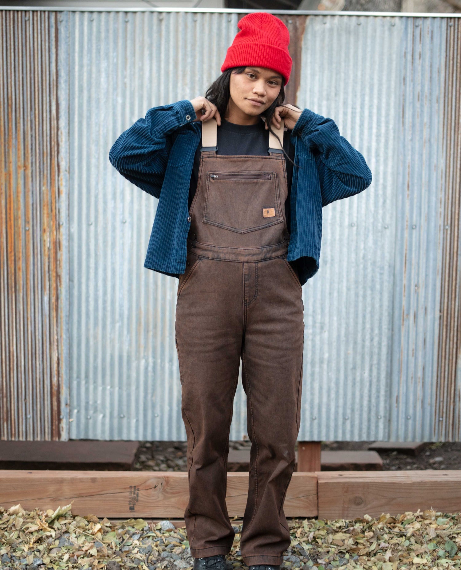 Person wearing brown overalls and a red beanie standing against a corrugated metal wall.